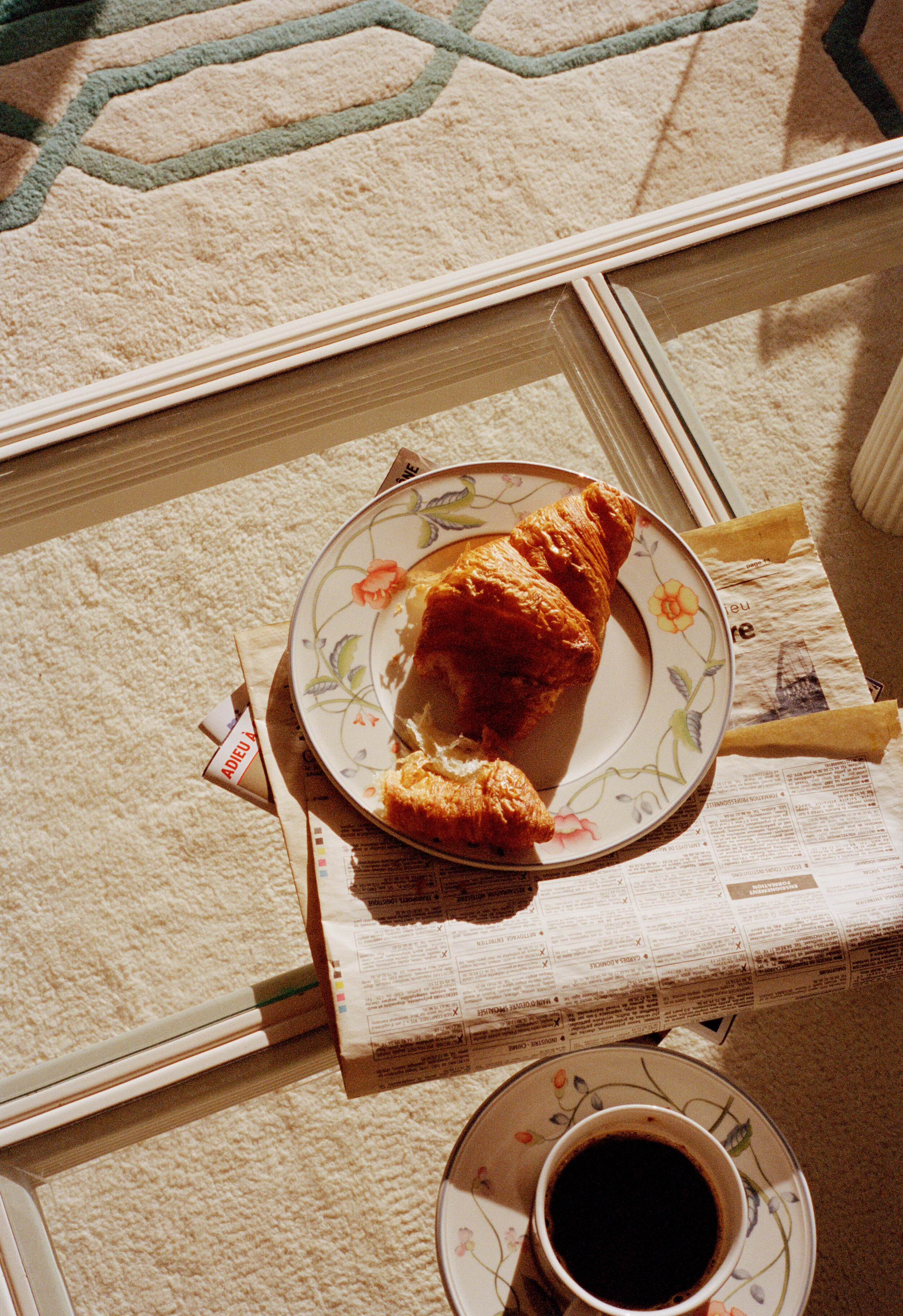 A breakfast scene with a plate of croissants, a cup of black coffee, and newspapers on a glass table near a window, with sunlight streaming in.