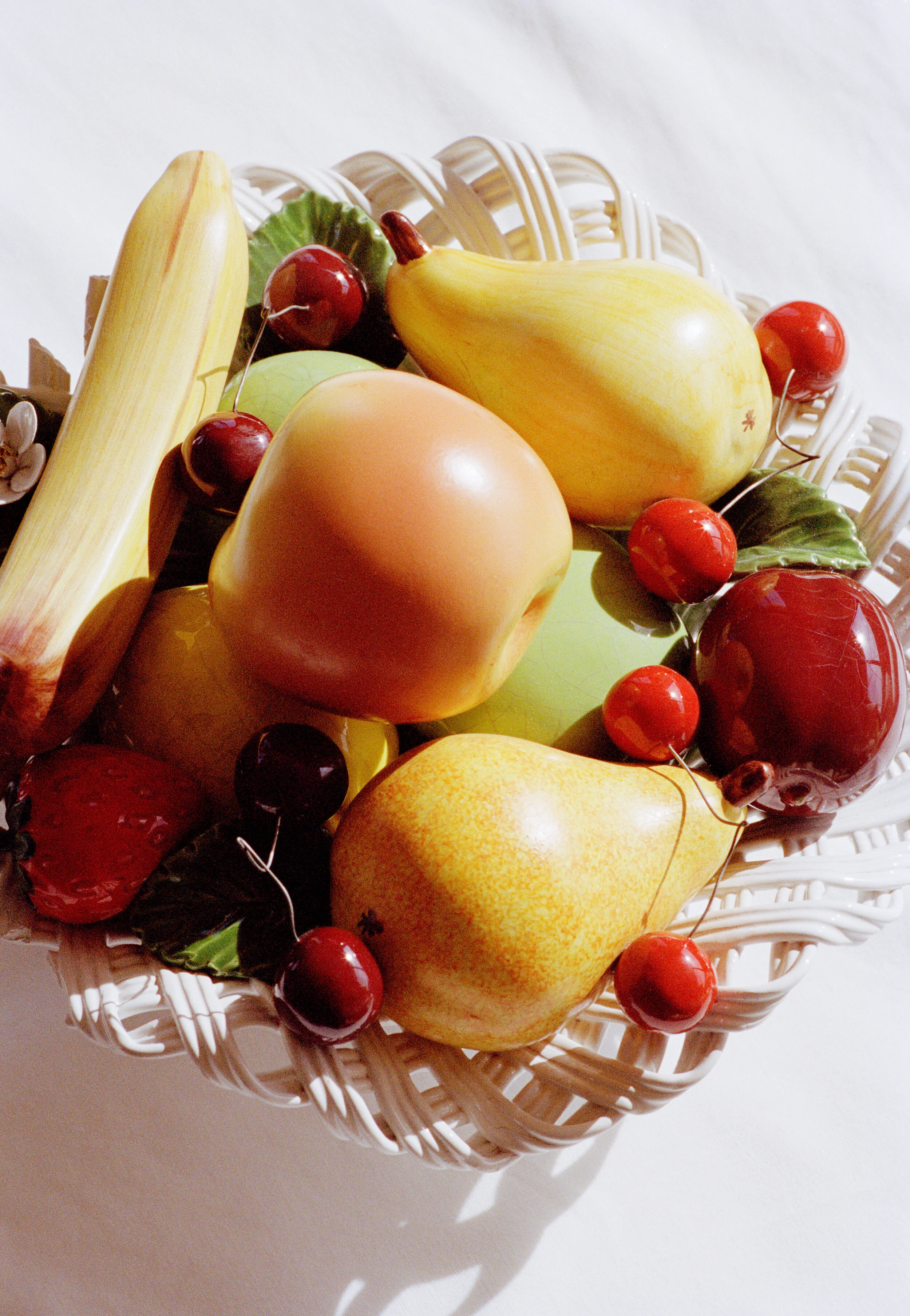 A white wicker basket filled with artificial pears, apples, cherries, and leaves on a white surface.