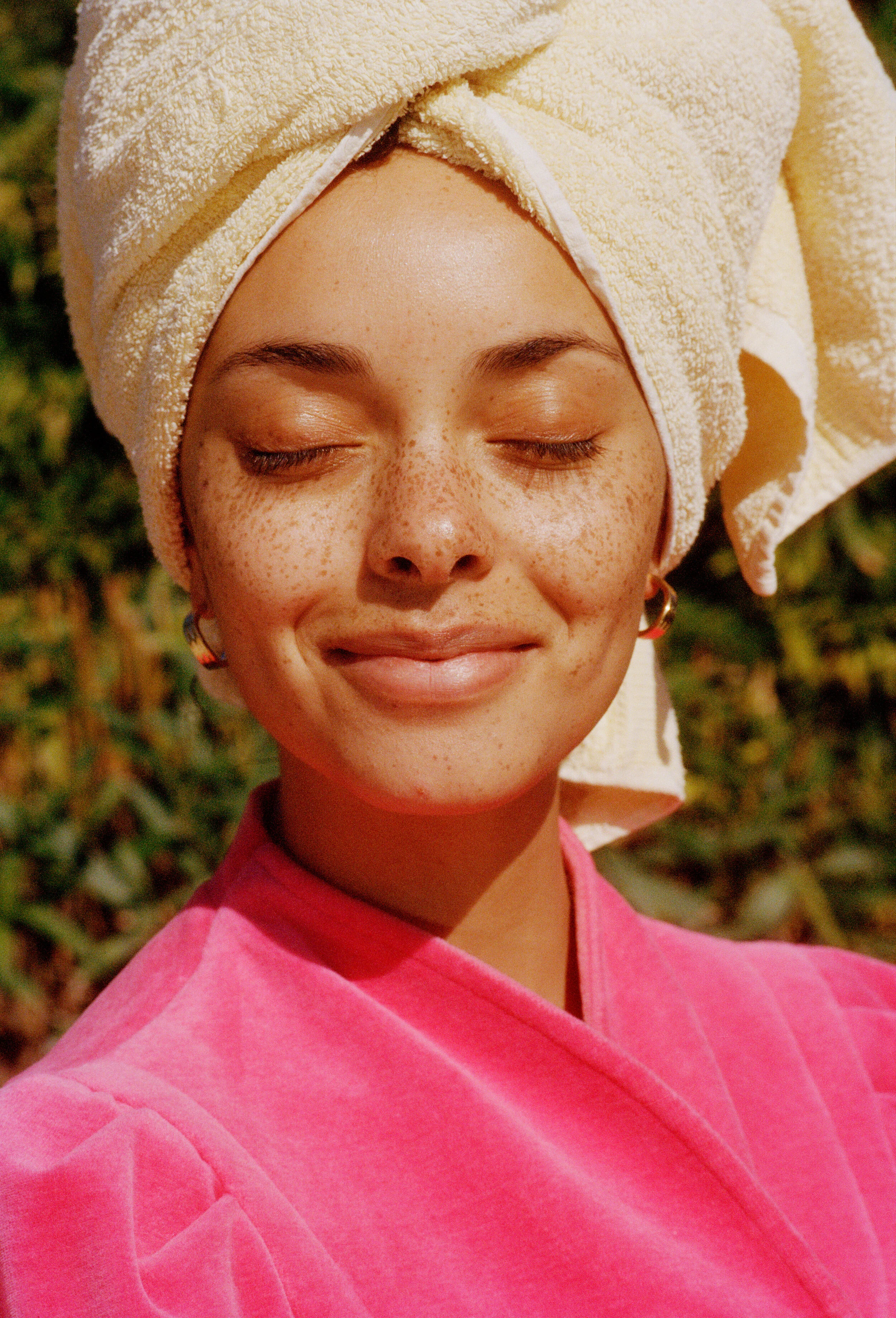 A young woman with a towel wrapped around her head, wearing a pink bathrobe, smiling peacefully with closed eyes, outdoors with greenery in the background.