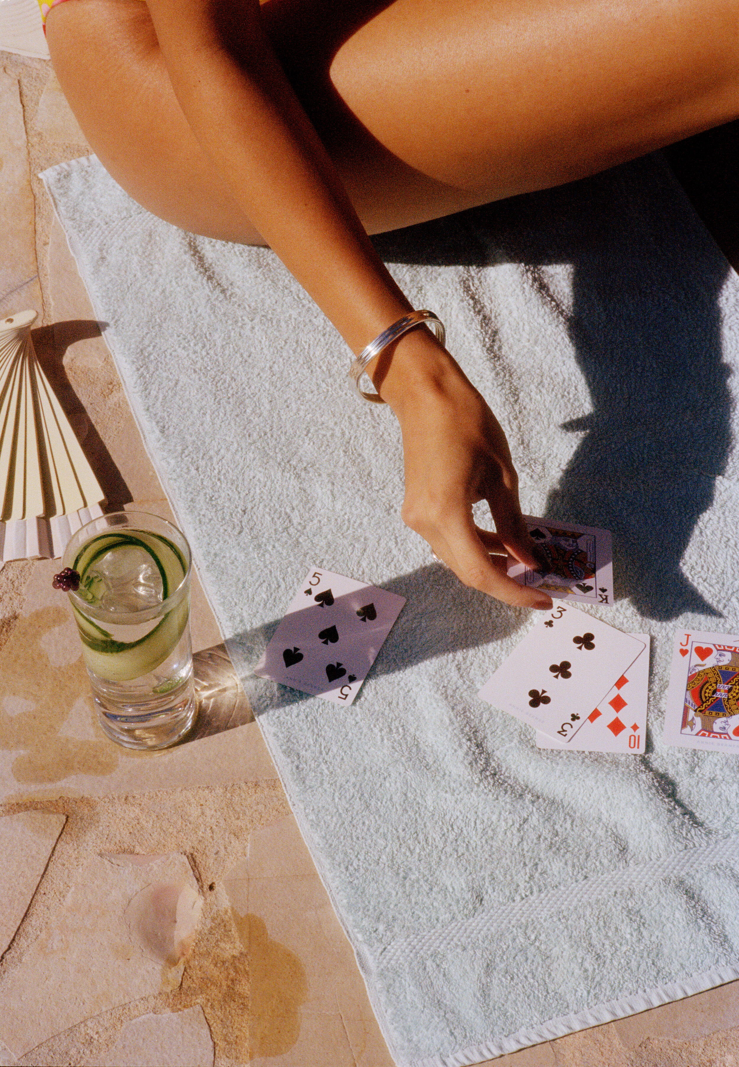 A woman sitting on a towel outdoors, playing cards, with a drink containing cucumber and a cherry nearby.