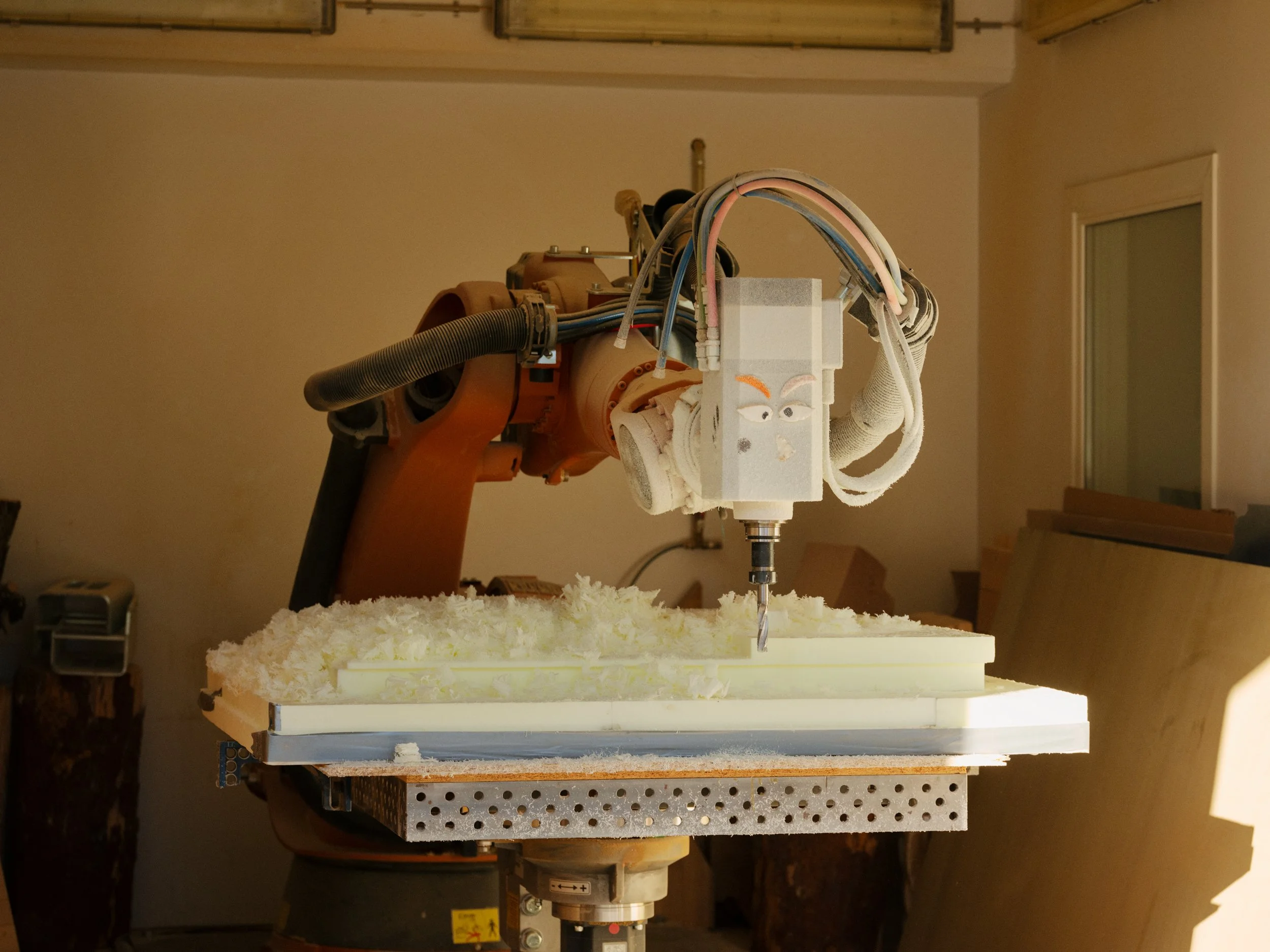 A robotic arm with a face design on a white box, holding a drill, working on a material with wood shavings around, in a workshop.
