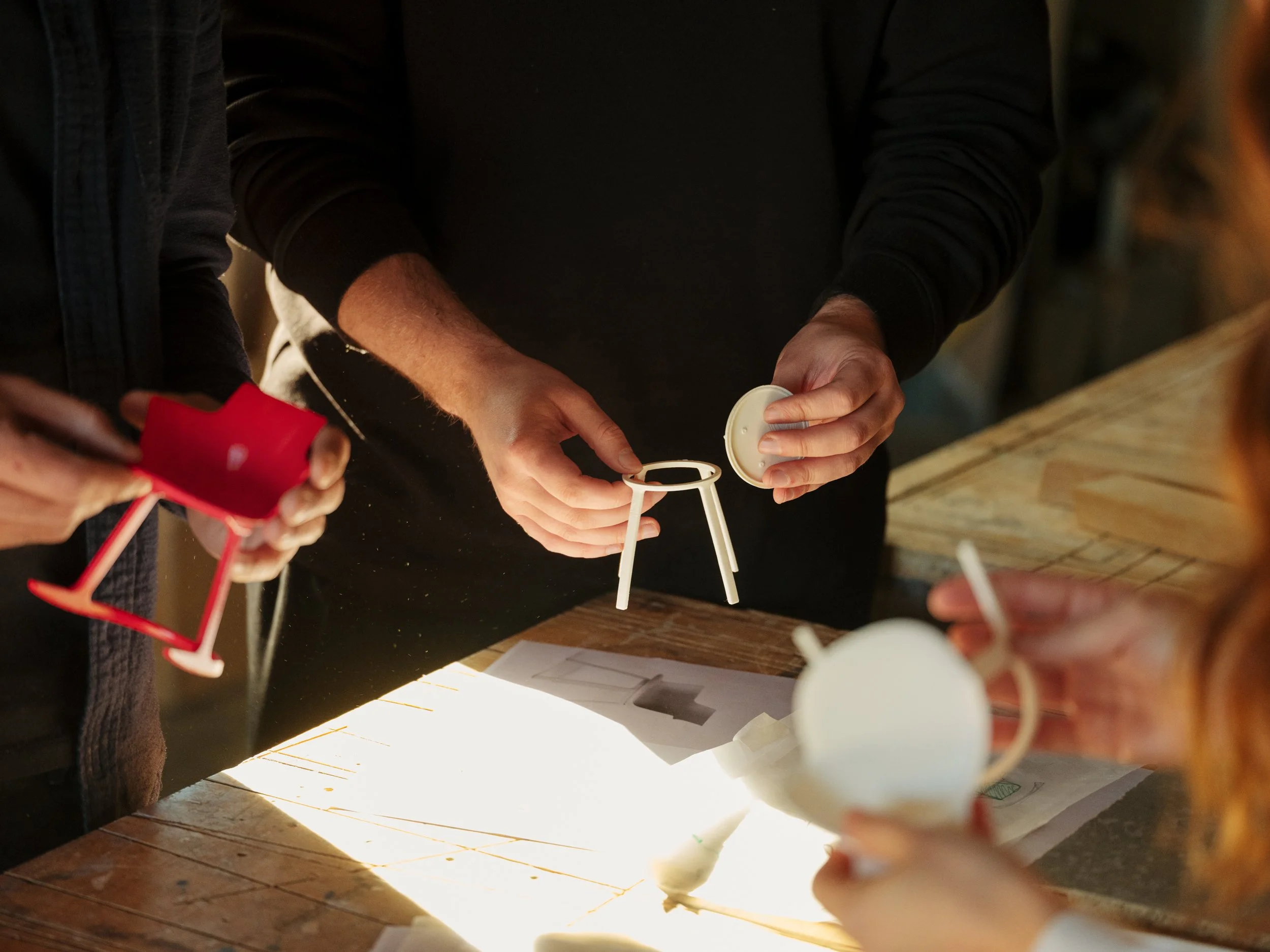 People assembling and inspecting small tea kettles and cups on a wooden table under a lighted surface.