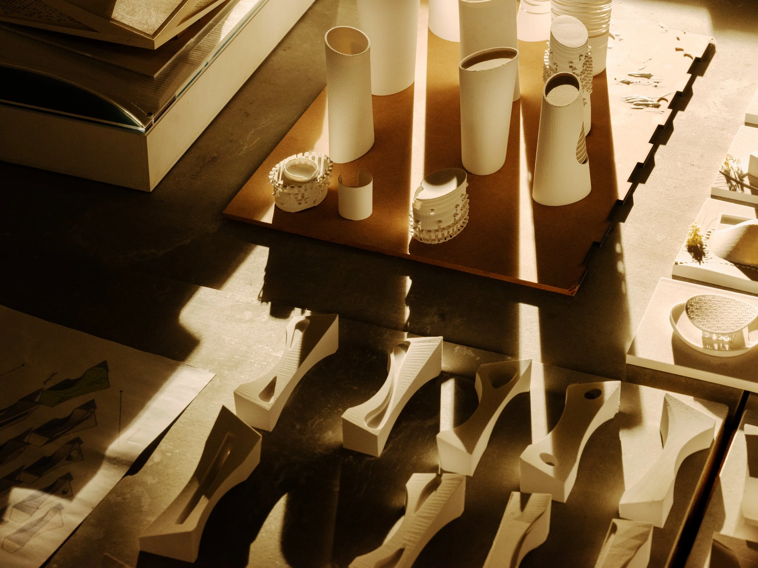 Architectural model components and sketches displayed on a table, including white sculptural elements, cylindrical structures, and paper drawings, with warm lighting casting shadows.