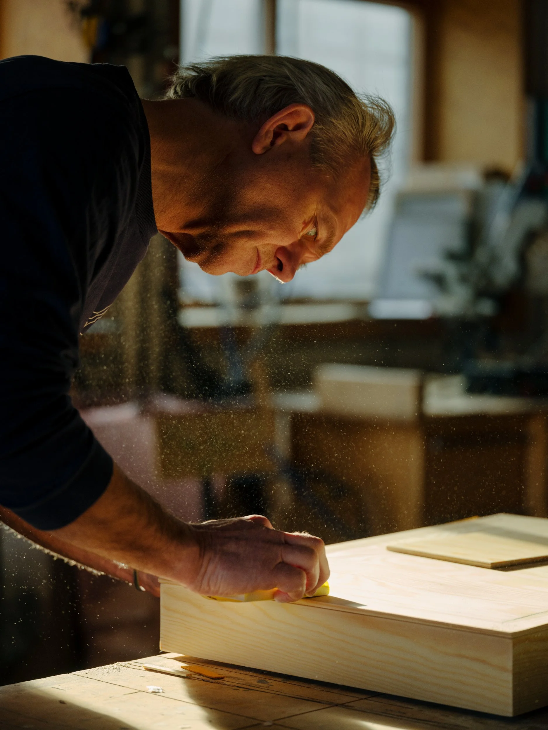 A man working on a piece of wood in a woodworking shop, using a tool to smooth or shape the wood, with sawdust flying around.