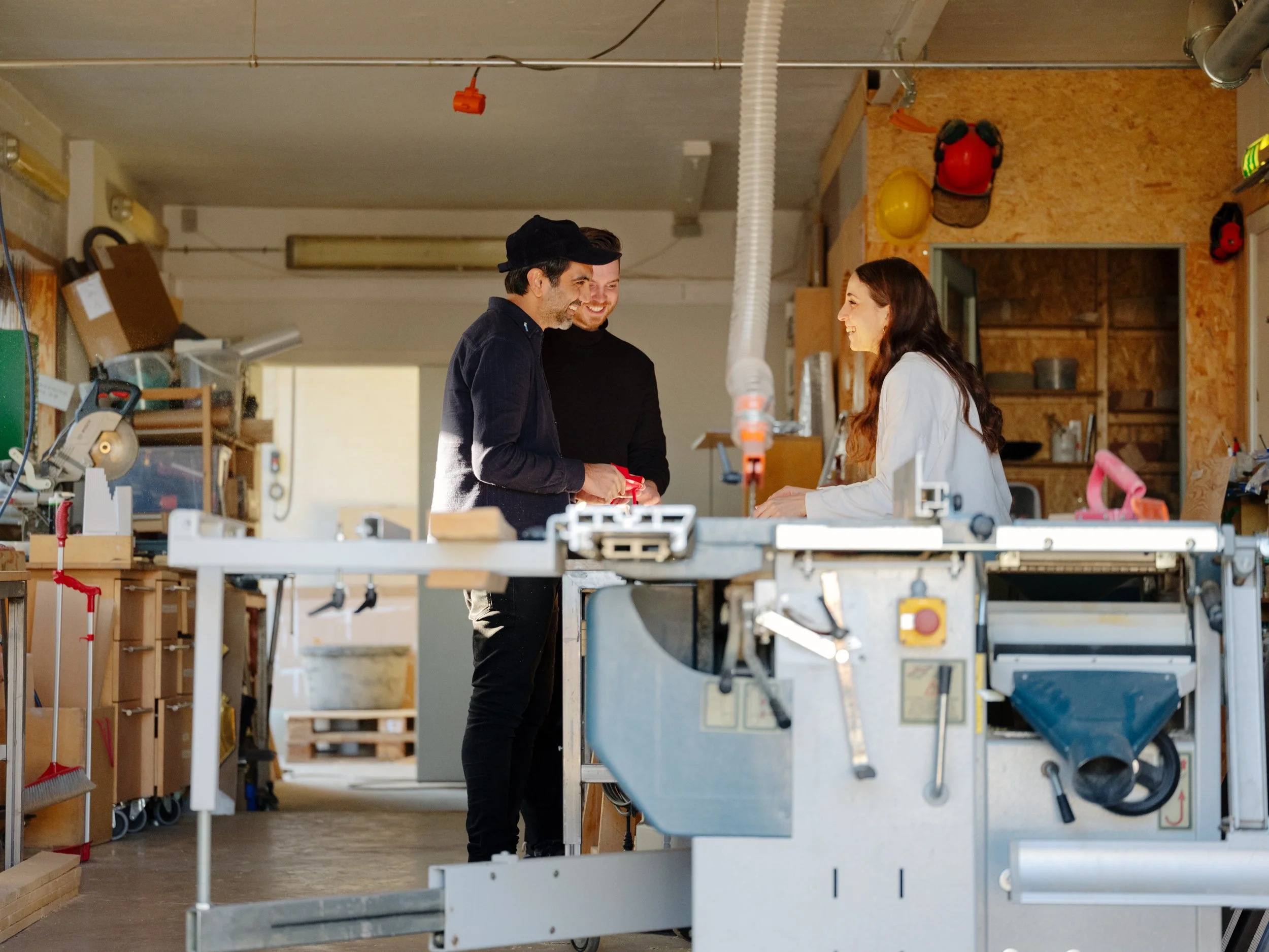 Three people inside a woodworking workshop, a woman on the right talking to two men on the left, one of whom is giving her a small red object, with woodworking tools and equipment in the background.