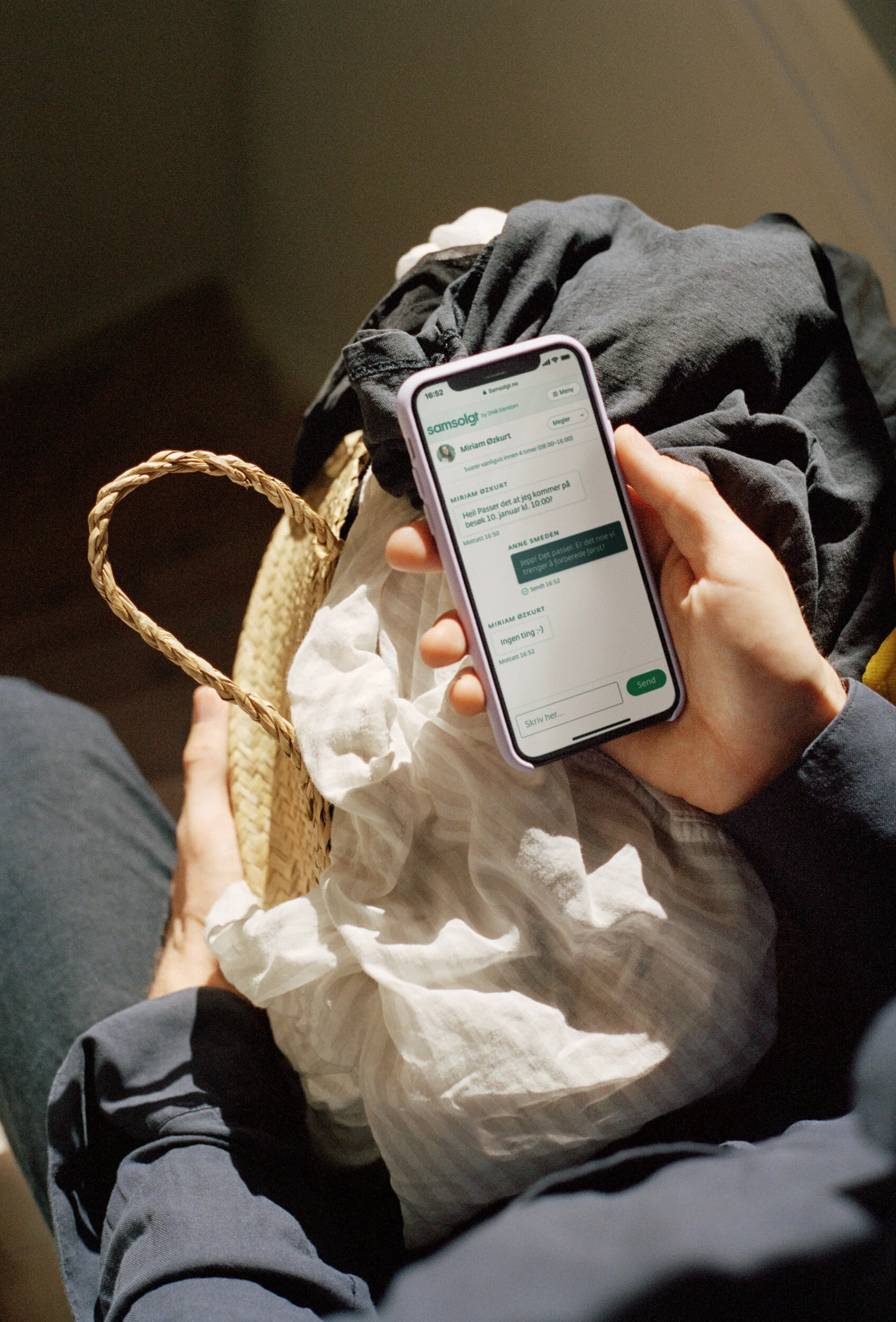 Person holding smartphone with messaging app, sitting on a bench with a woven purse and clothing items around.