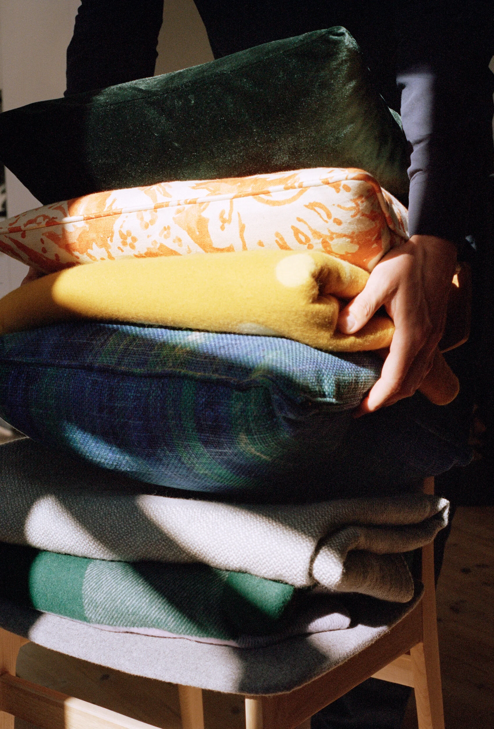 Person stacking folded clothes on a chair, including black, orange patterned, yellow, plaid, checked, and beige fabric.