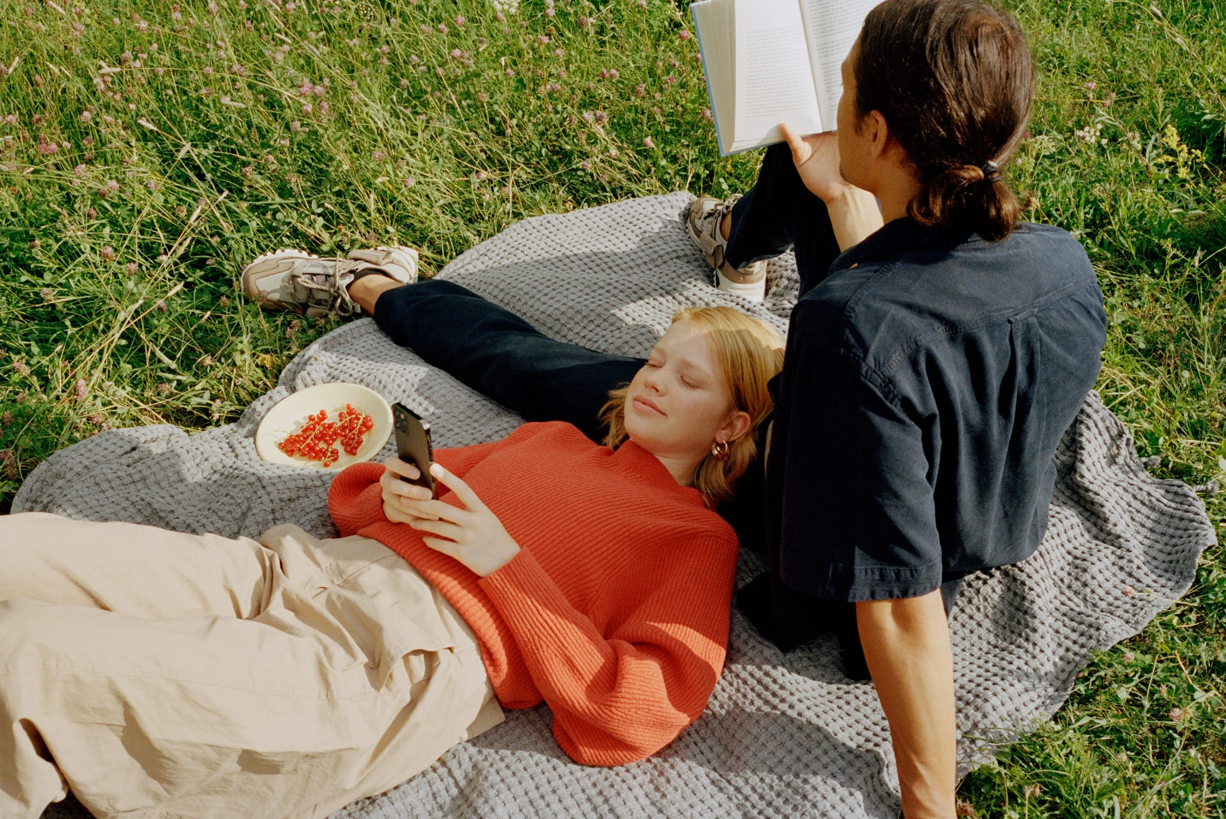 Two women relaxing outdoors on a blanket in a grassy field, one lying down looking at her phone, the other sitting up reading a book, with a bowl of red berries nearby.