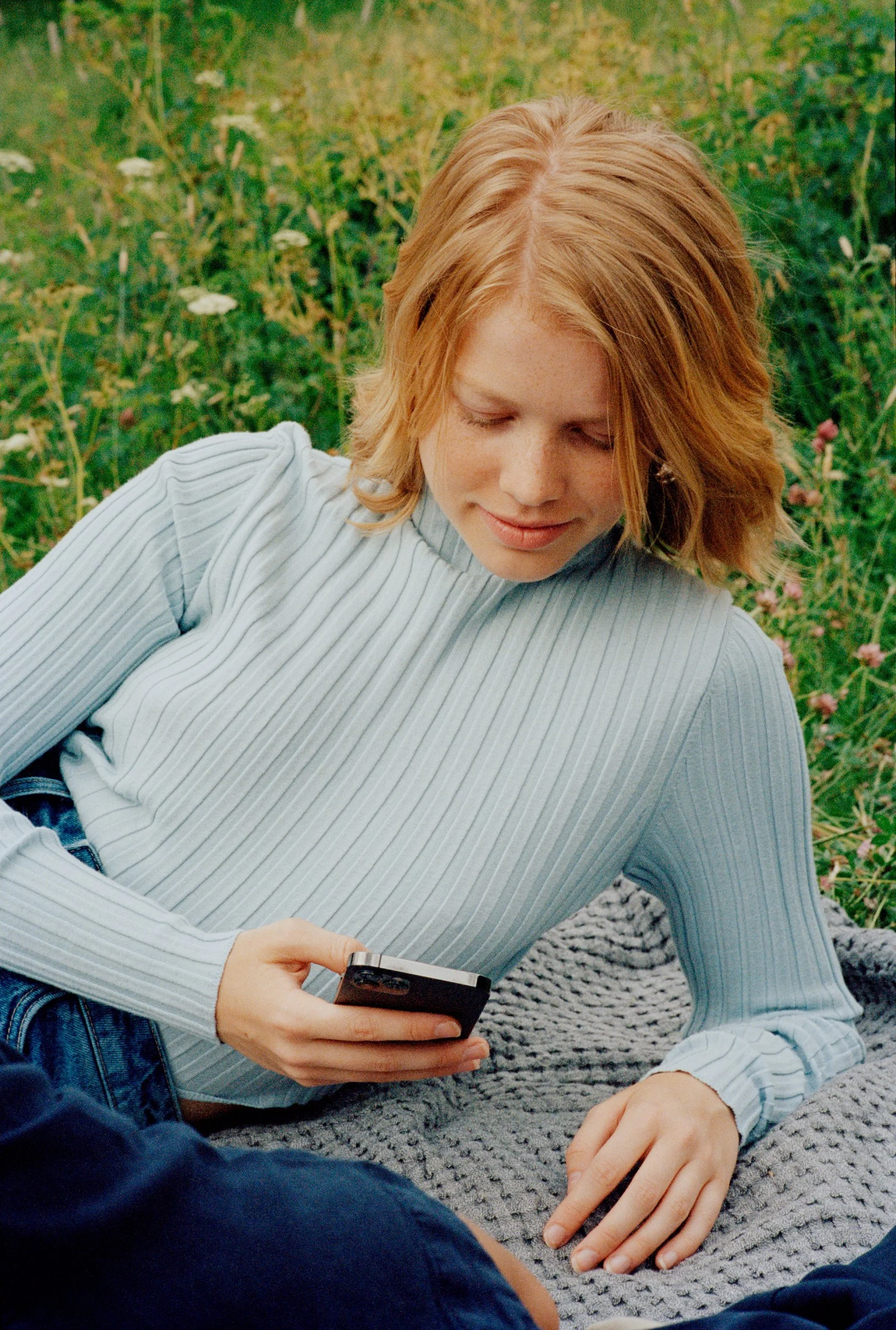 A young woman with red hair and freckles lying on a gray blanket outdoors in a green field, looking at her phone.