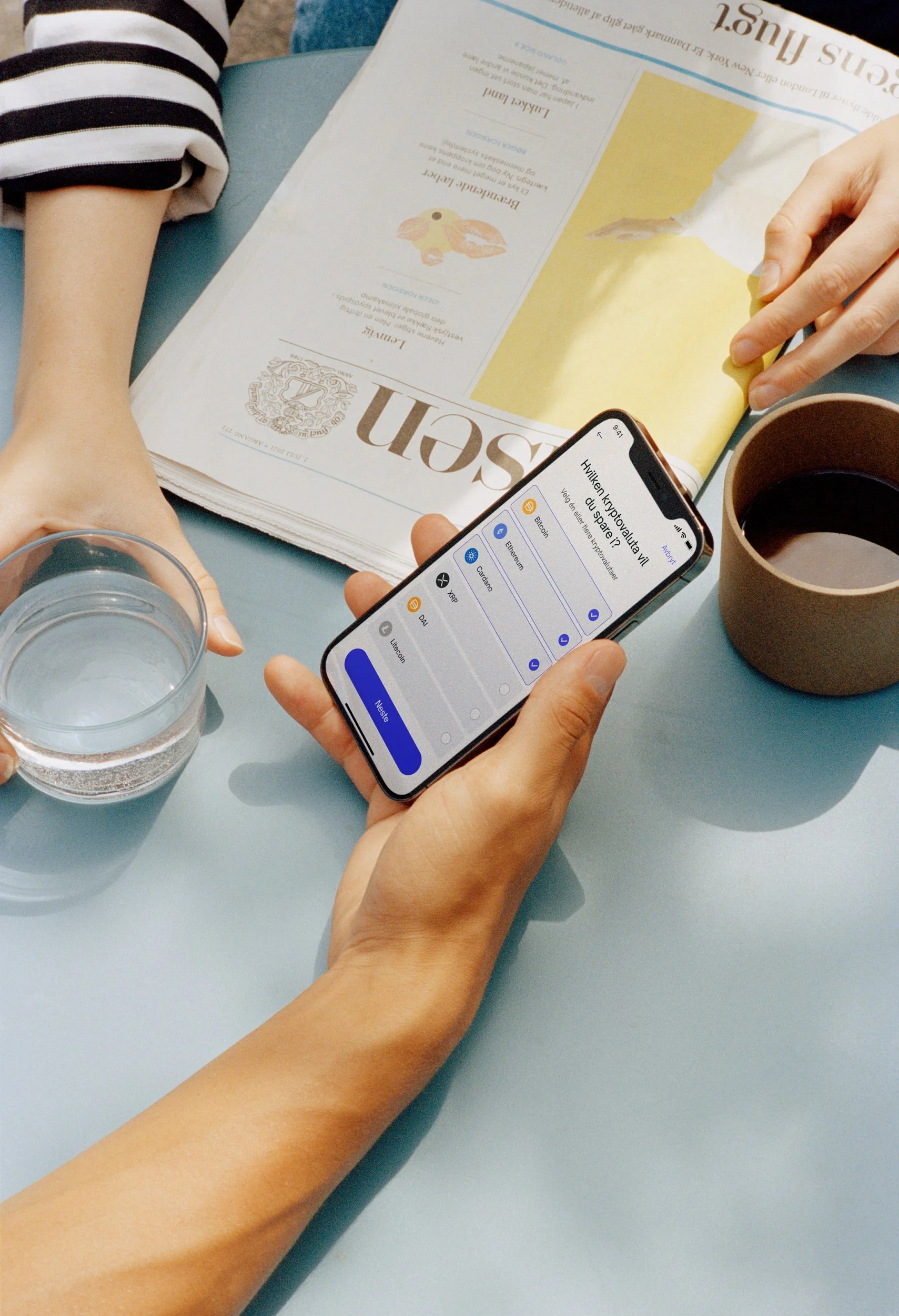 Person holding a smartphone displaying a cryptocurrency quiz app, with a glass of water, a cup of coffee, a yellow book, and a newspaper on a blue table.