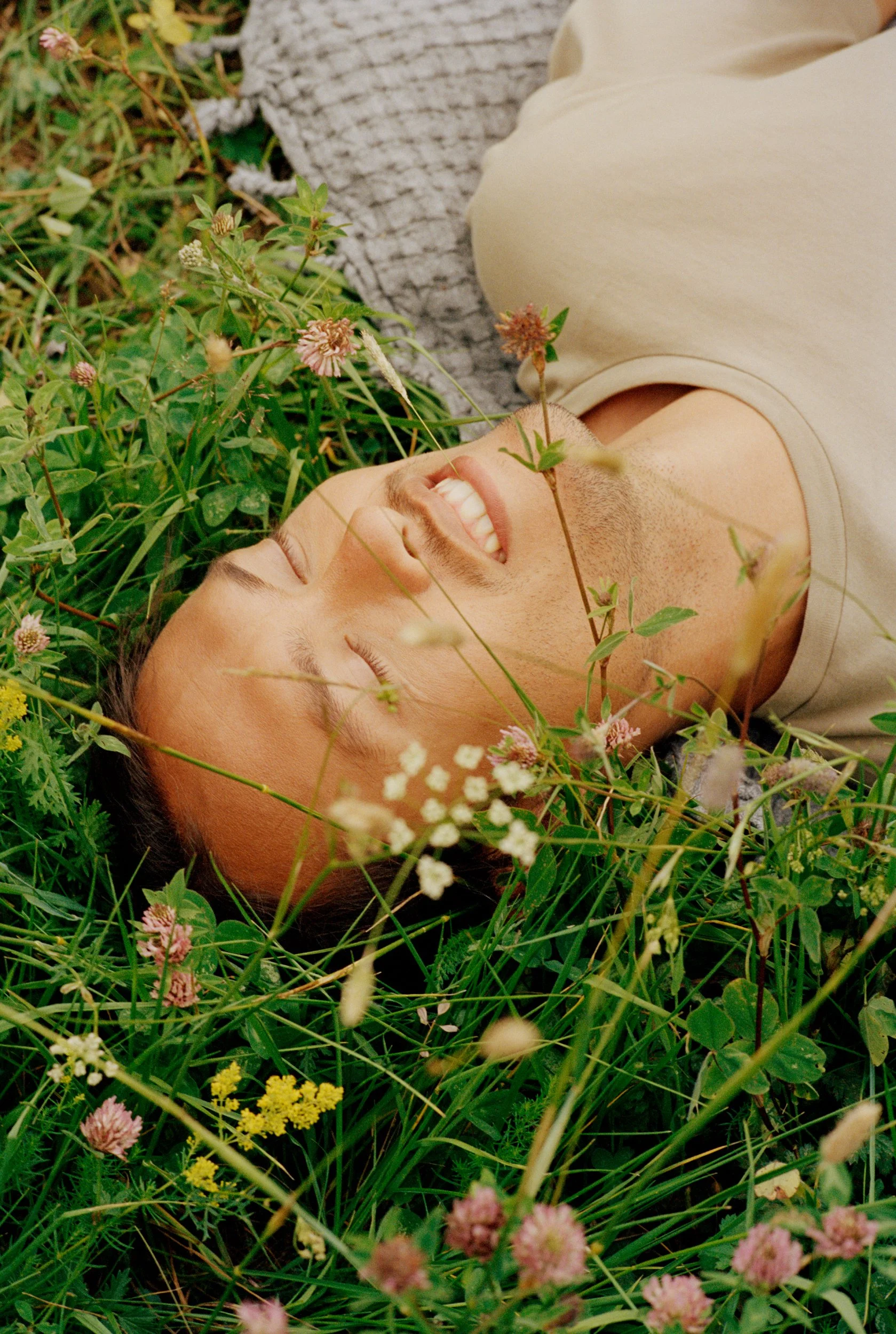 A young man lying on his back in a field of green grass and wildflowers, smiling with eyes closed.