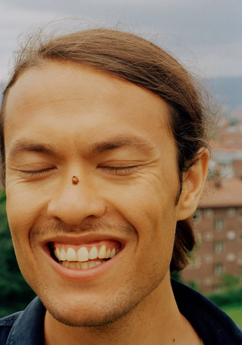 A young man with light skin and brown hair is smiling with eyes closed, outdoors during daytime, with a blurred cityscape in the background.