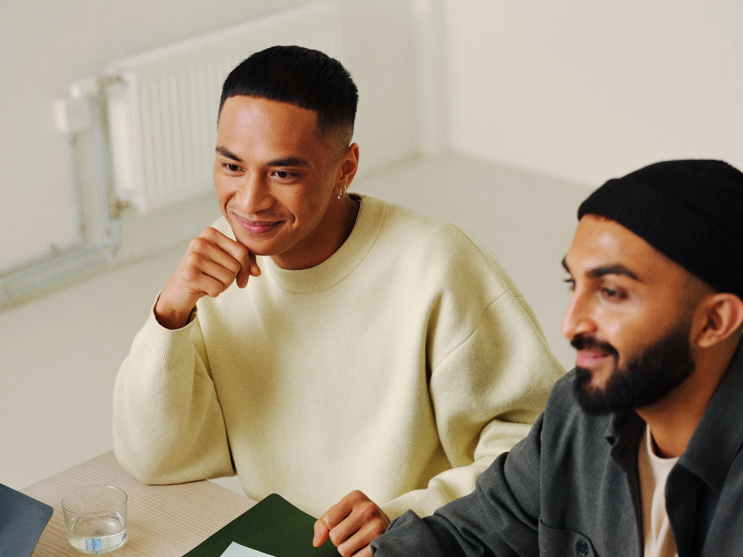 Two men sitting at a table during a meeting, one wearing a cream-colored sweater and the other in a black beanie and gray jacket, with a glass of water and a notepad on the table.