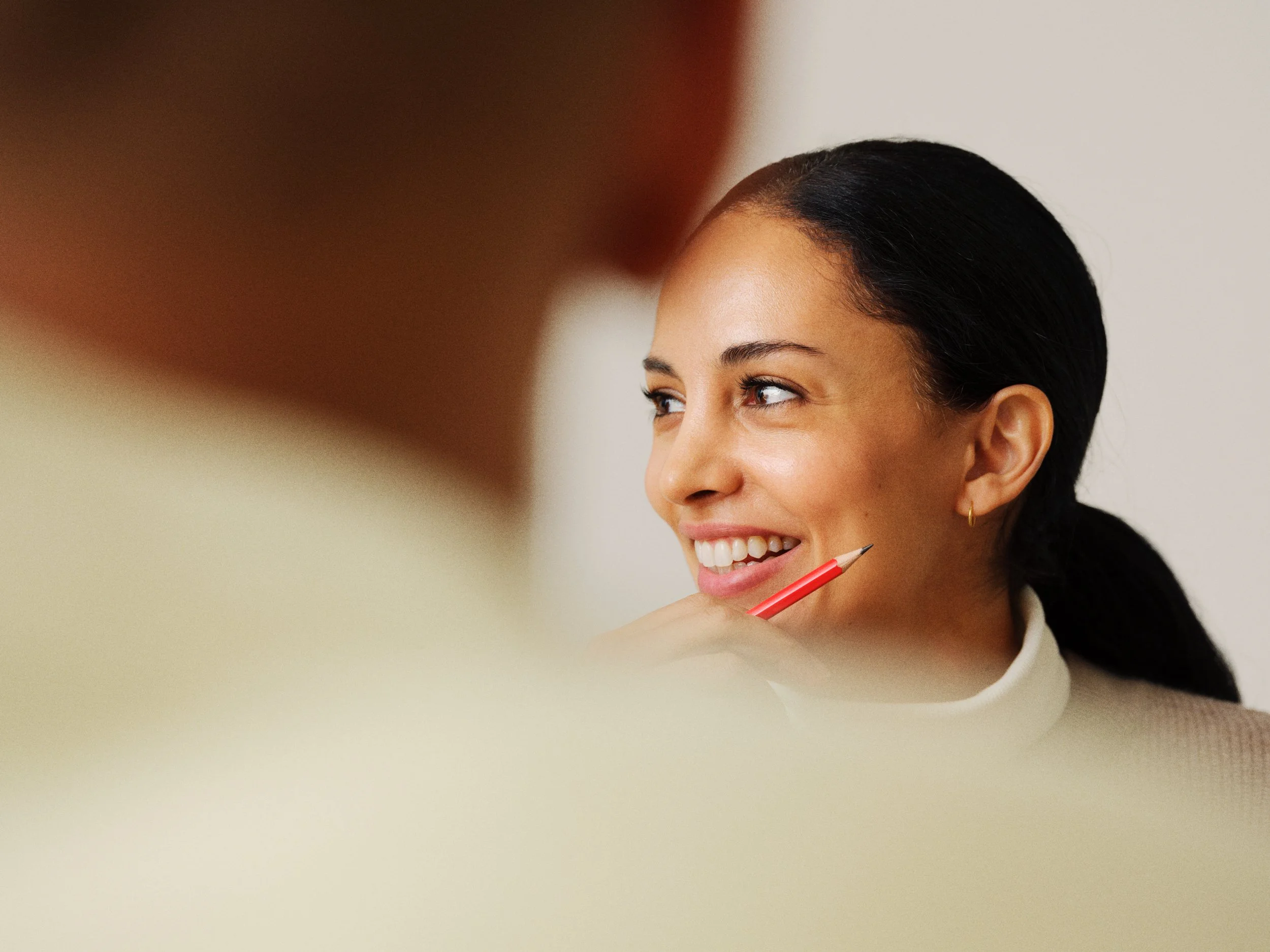 A woman with dark hair pulled back, smiling and holding a red pencil near her chin, listening to someone in the foreground, partially visible.