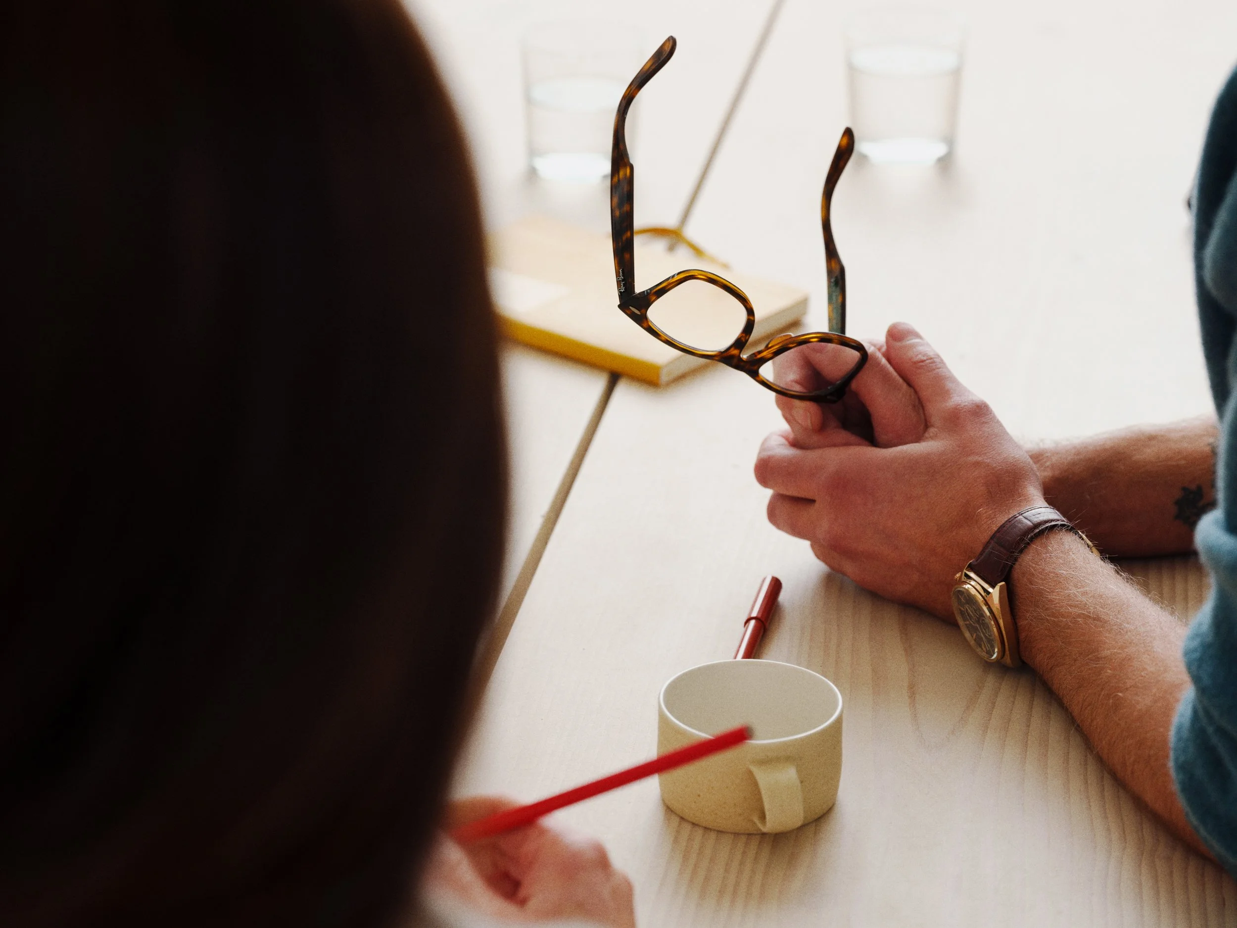 Person holding tortoise shell eyeglasses on a light wooden table, with a white mug, red pen, and glasses in the background.
