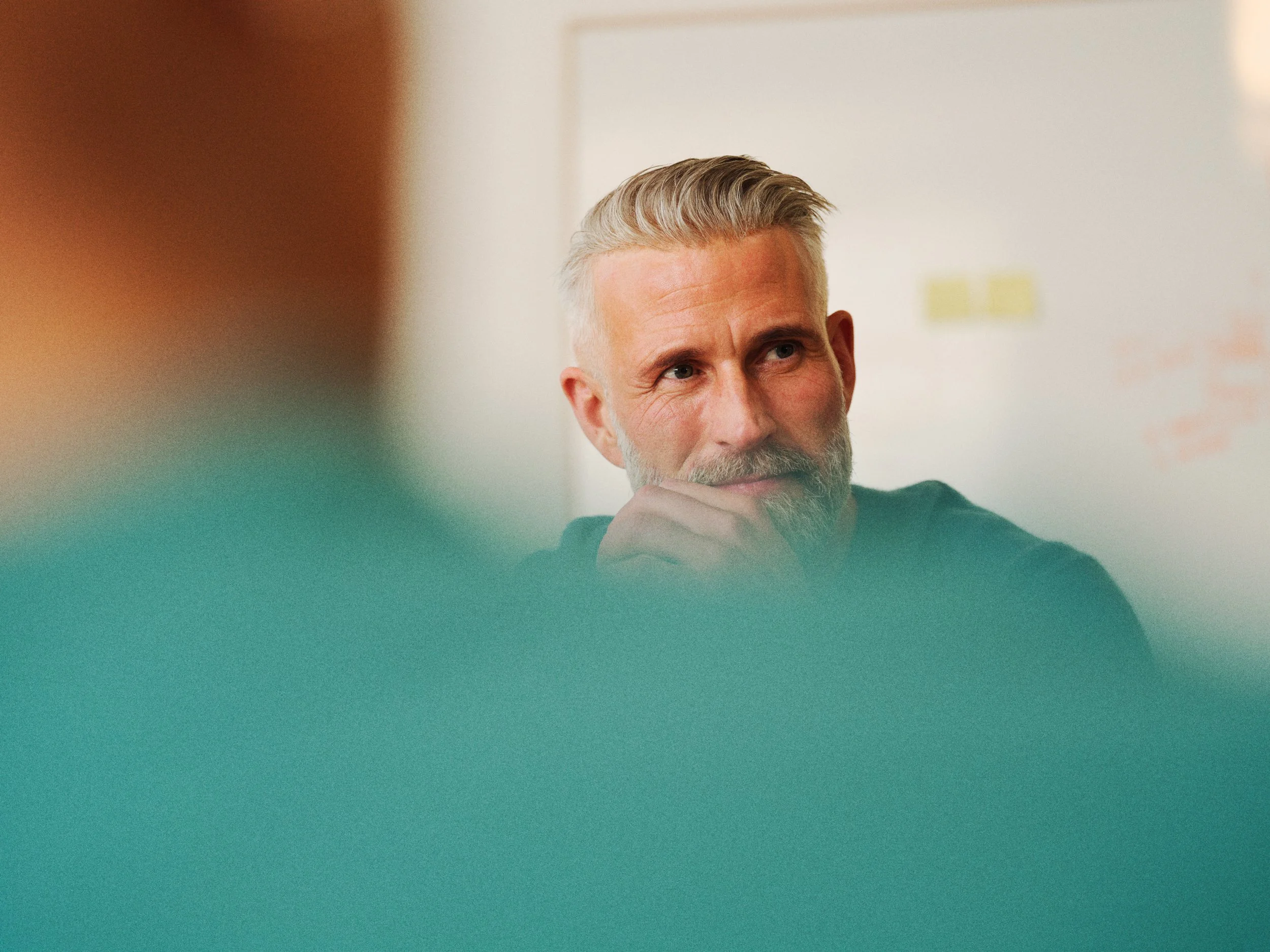 A middle-aged man with gray hair and a beard, sitting thoughtfully in a meeting room, with out-of-focus colorful objects in the foreground.