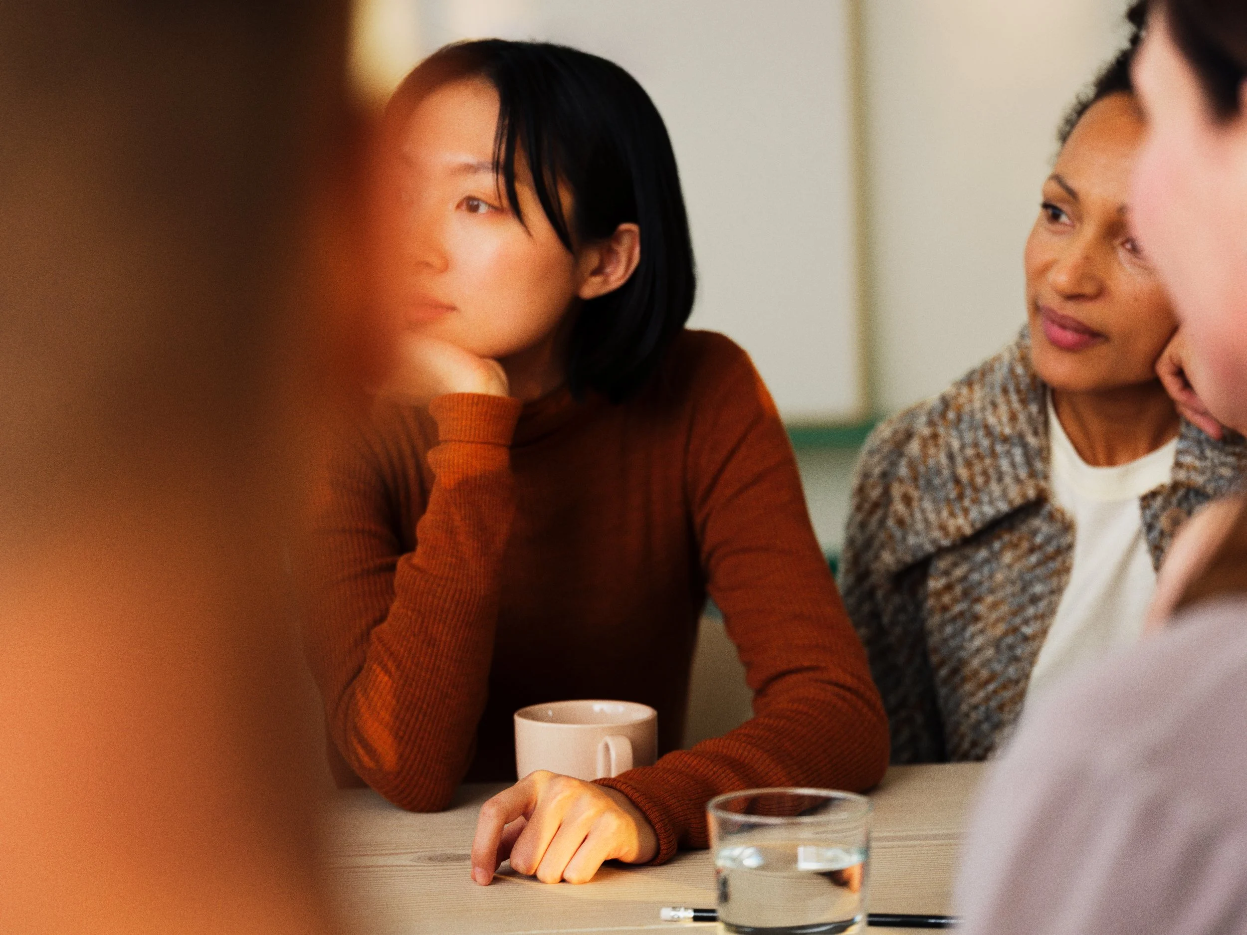 A group of diverse women engaged in a serious discussion around a table, with one woman resting her chin on her hand.