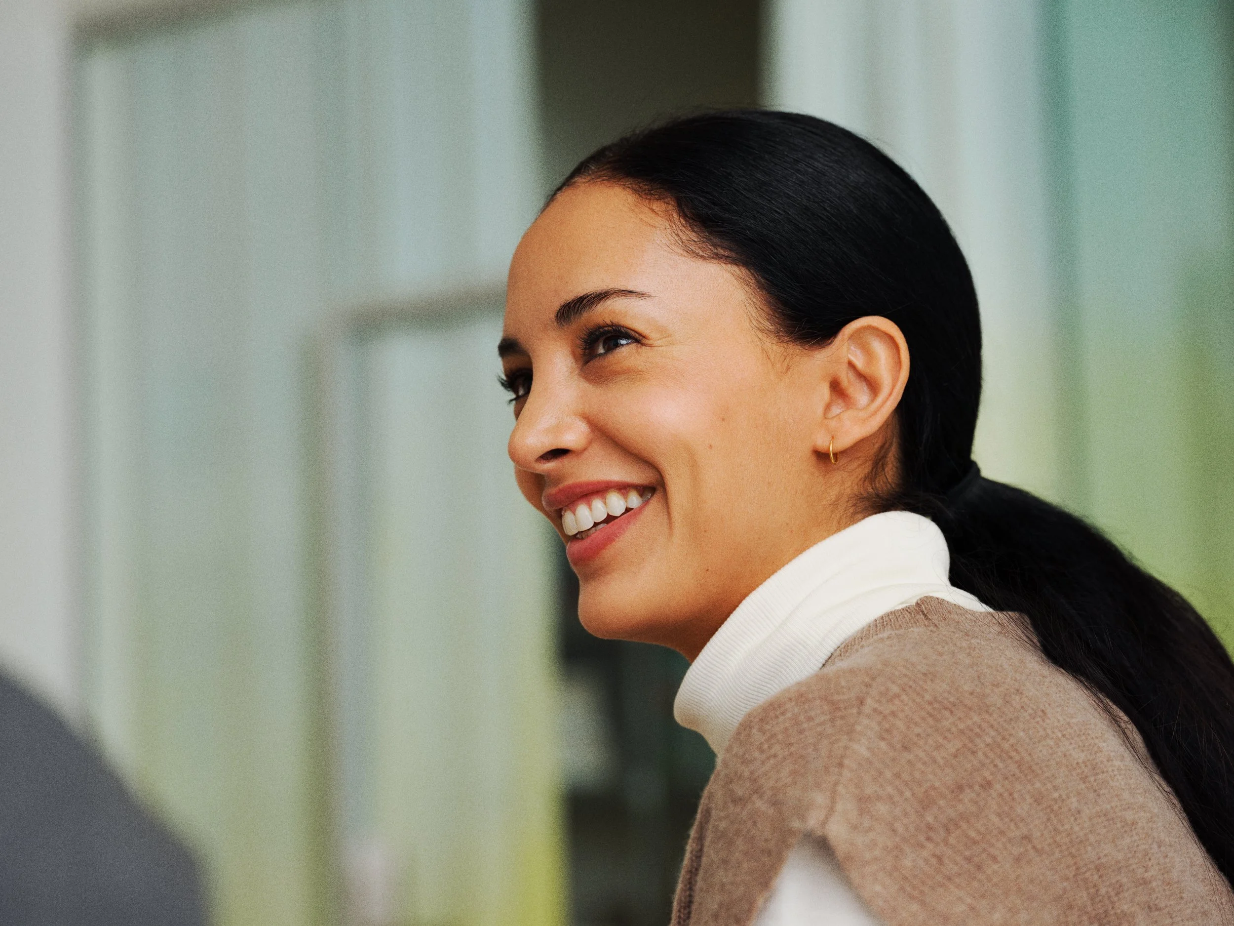 A woman with black hair, smiling, and wearing a beige sweater with a white turtleneck, sitting in a well-lit room.