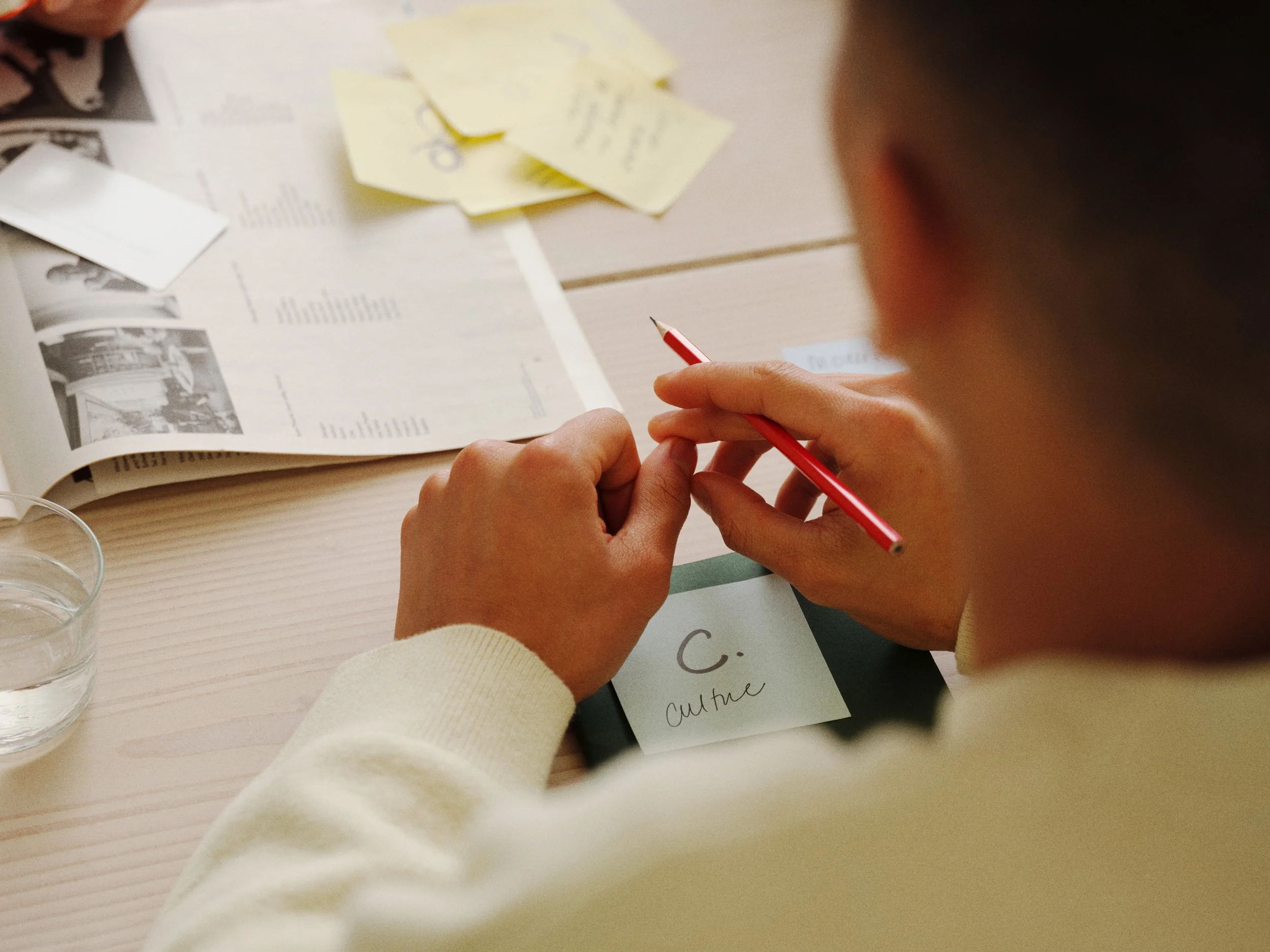 Person writing on sticky note at desk with papers, newspaper, and glass of water.