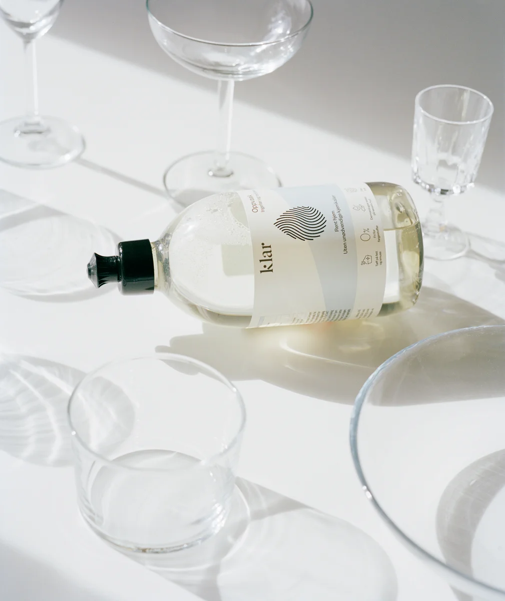 A bottle of J. Lair sparkling water lying on a white surface surrounded by transparent glassware, including wine glasses, a tumbler, and a bowl, with sunlight creating shadows.