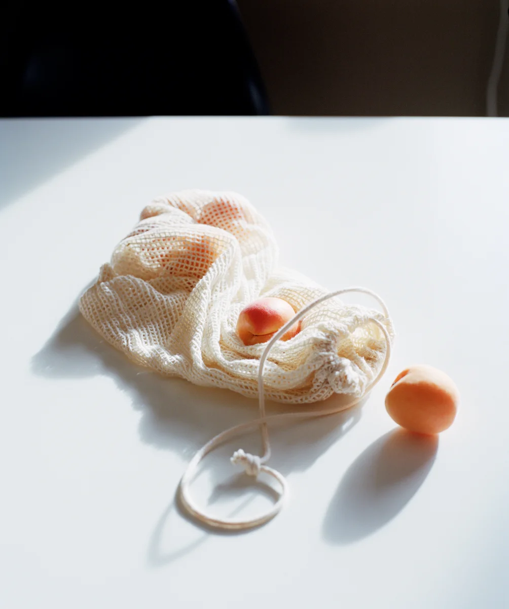 Peach fruits and a white mesh drawstring bag on a white surface with sunlight shadows.