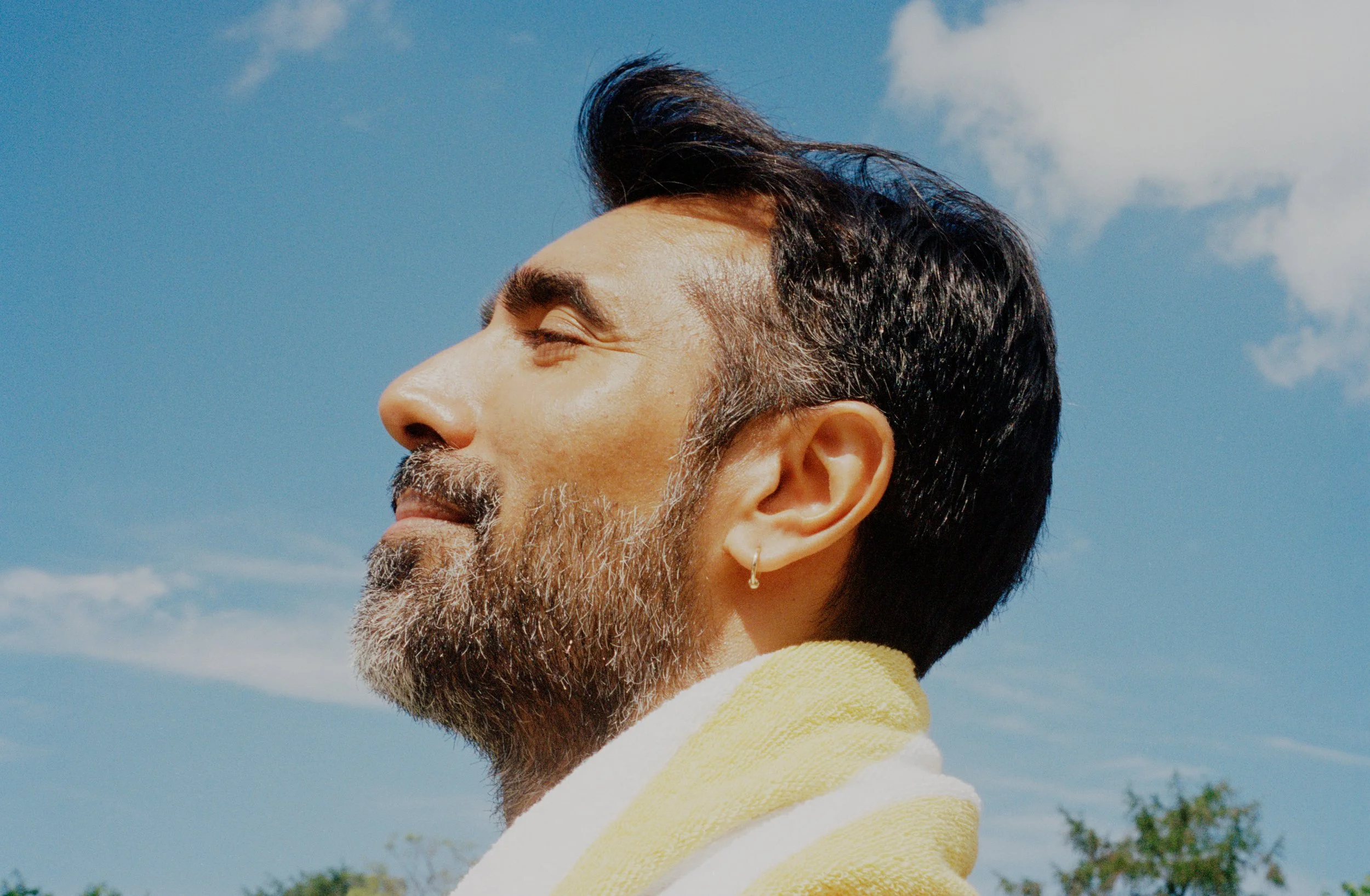 A man with a beard and mustache, wearing a yellow and white towel around his shoulders, enjoying the sunshine outdoors with eyes closed under a clear blue sky.
