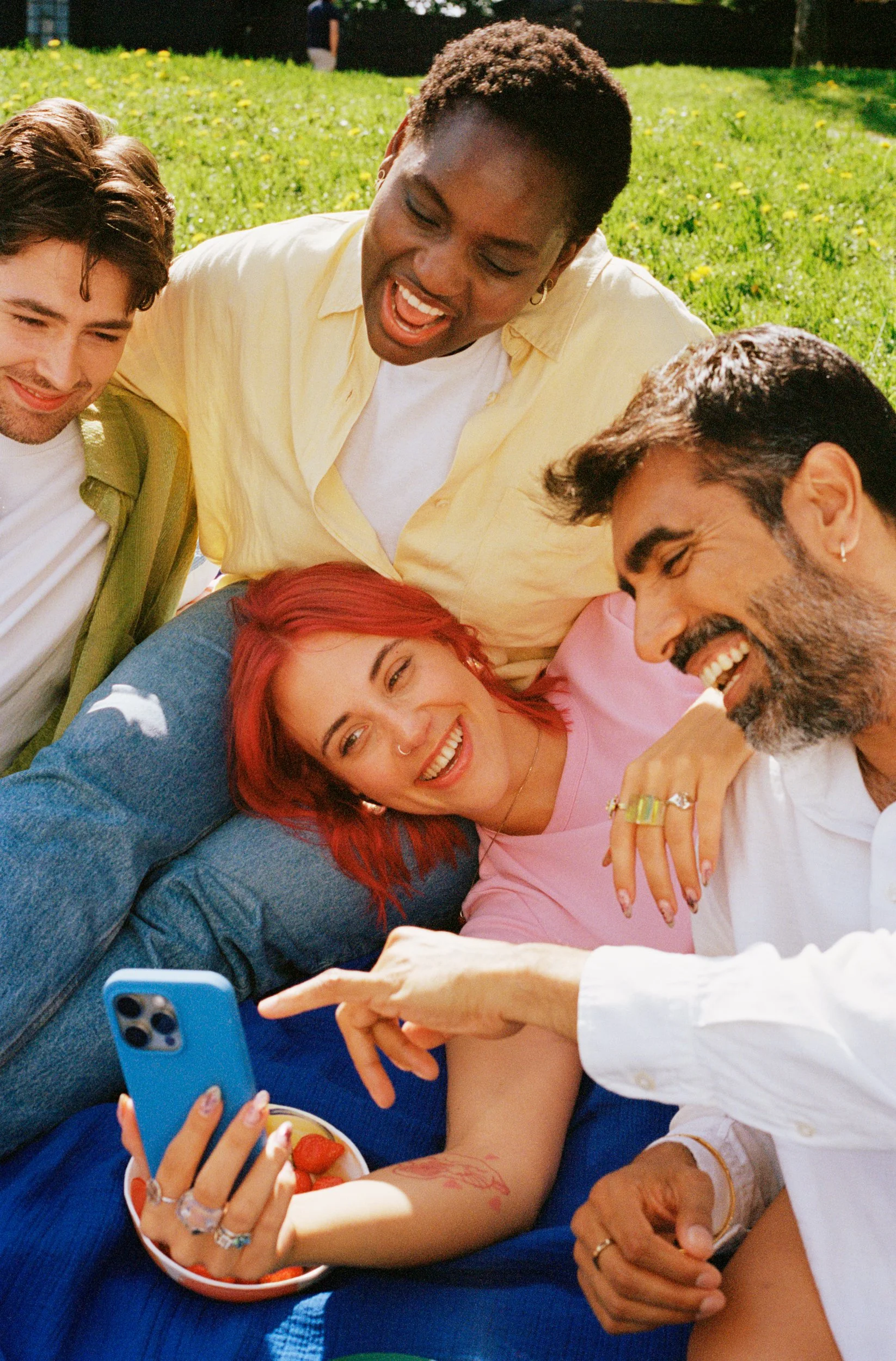 A group of five diverse friends lying on the grass and taking a selfie together, smiling and laughing.