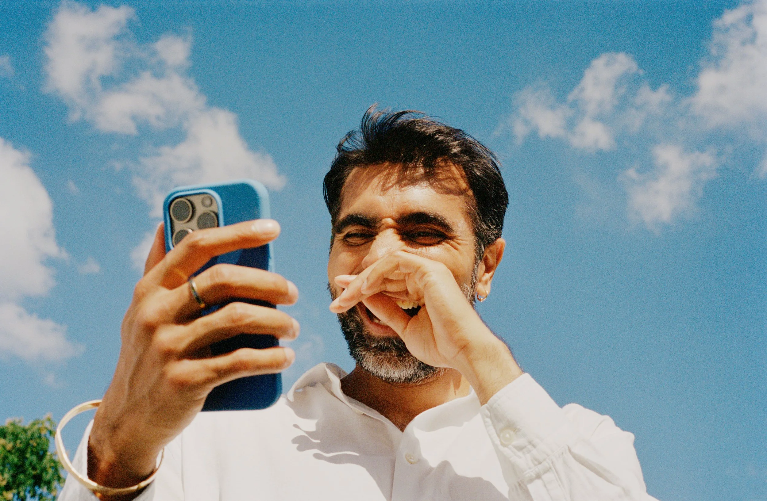 A man with dark hair and a beard, wearing a white shirt, is outdoors smiling and looking at his phone, with a blue sky and some clouds in the background.