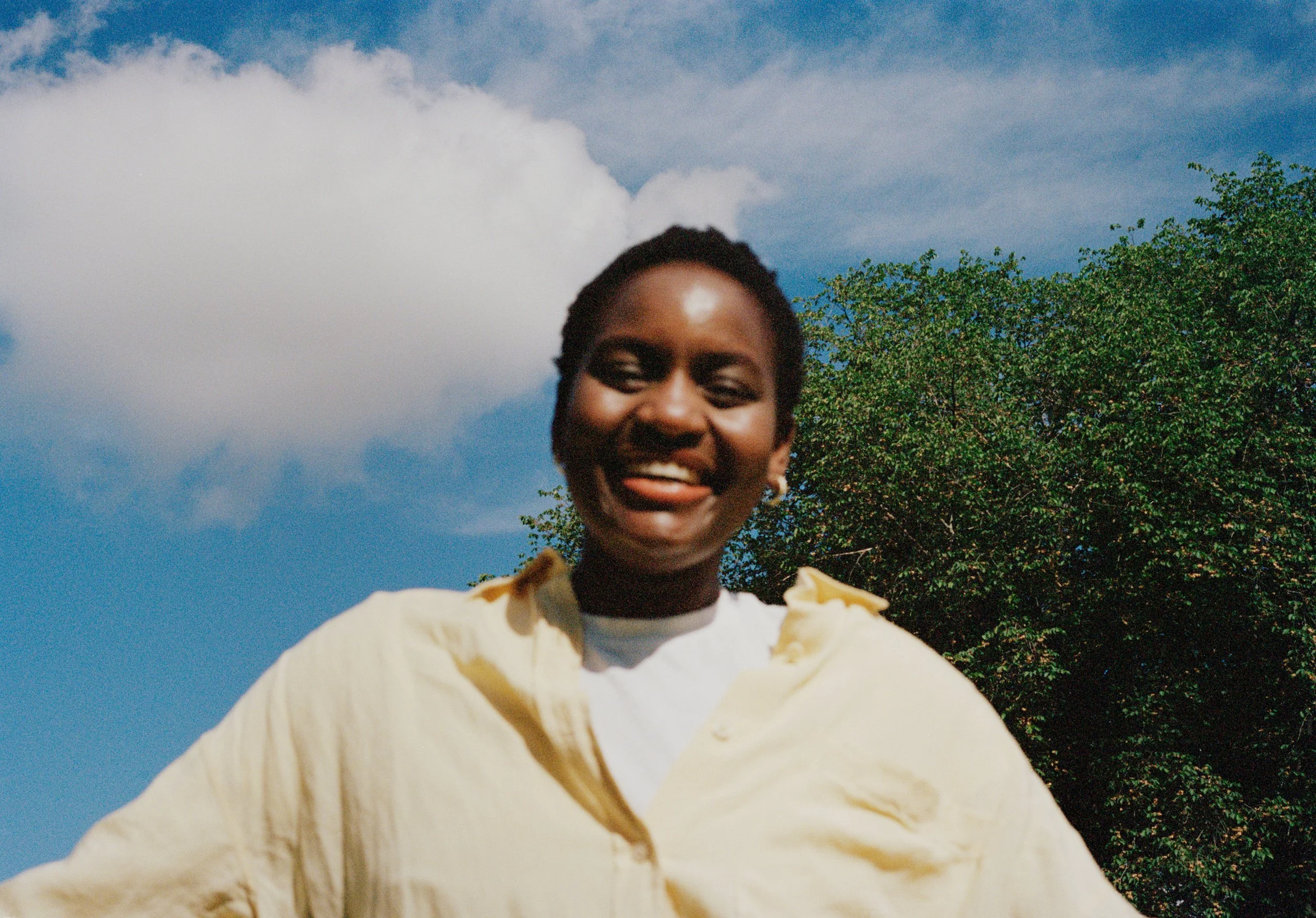 Smiling woman outdoors with a blue sky, white clouds, and green trees behind her.