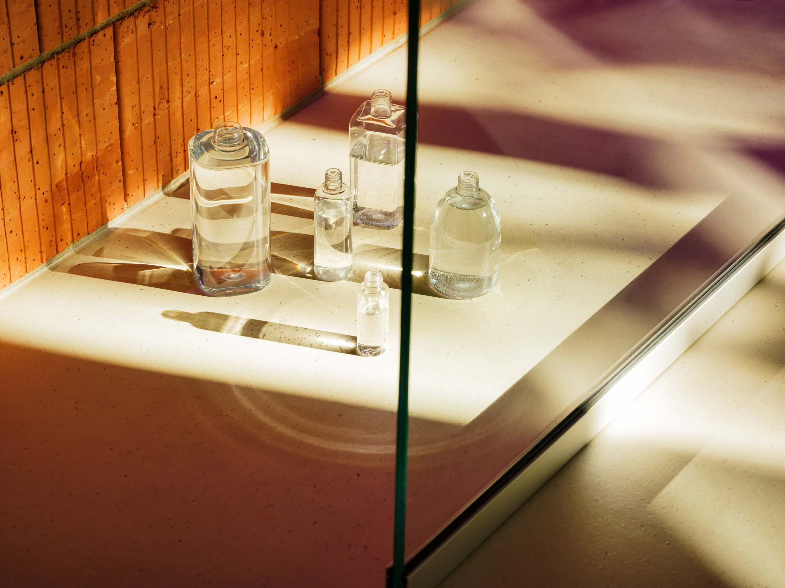 Various glass bottles filled with water on a white surface, reflected in a mirror, with a wooden wall behind and sunlight casting shadows.