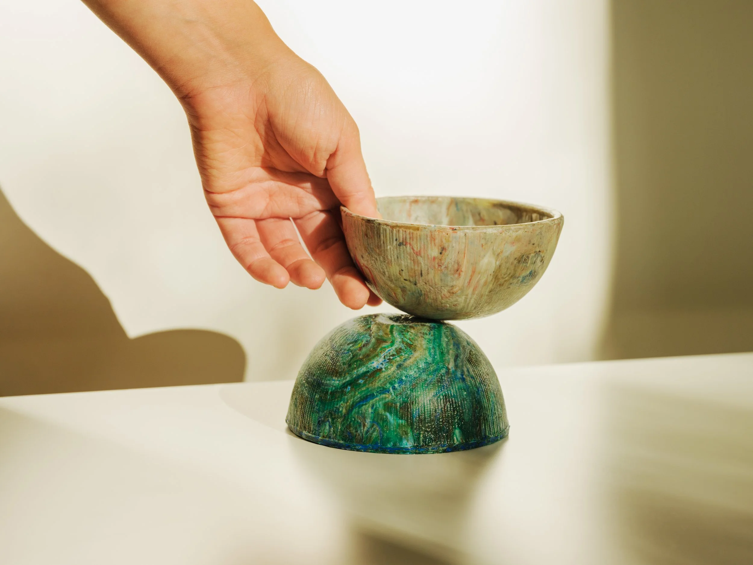 A hand carefully balances a ceramic bowl on top of a matching base, with a beige background.
