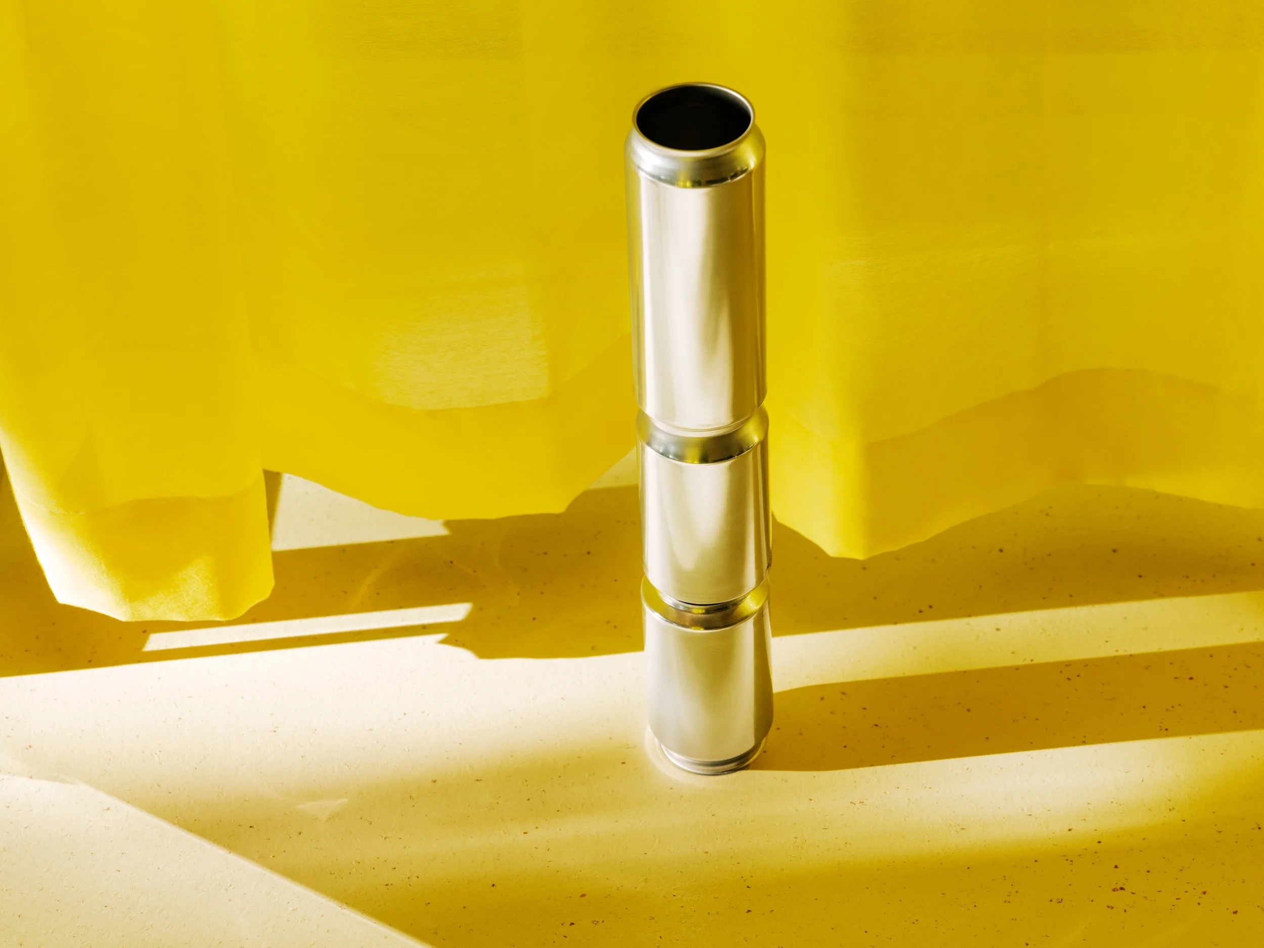 A metallic tube standing vertically on a light-colored space surface, with a yellow fabric background and shadows cast on the surface.