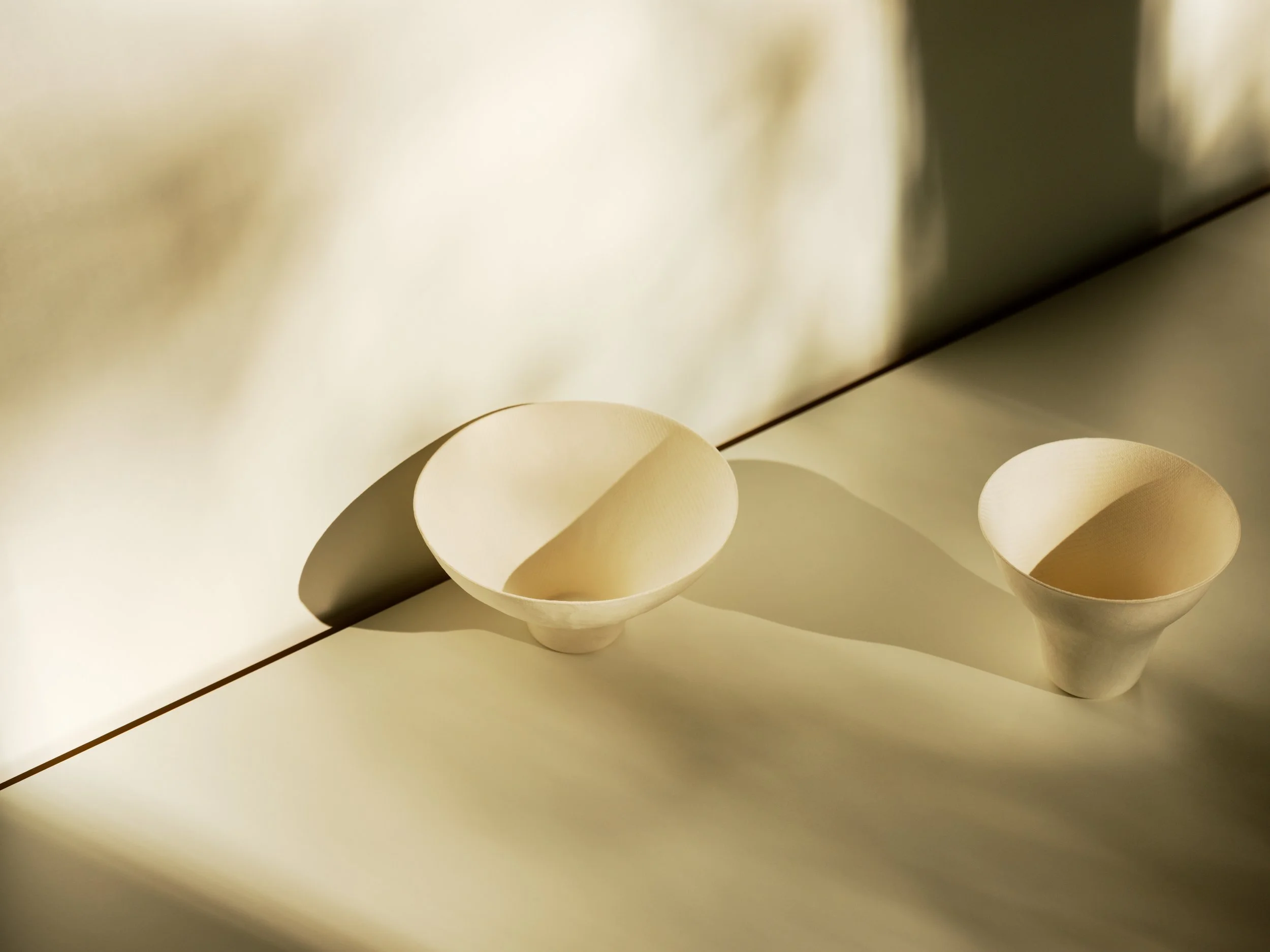 Two cream-colored ceramic bowls placed on a light surface against a cream-colored wall, casting elongated shadows.
