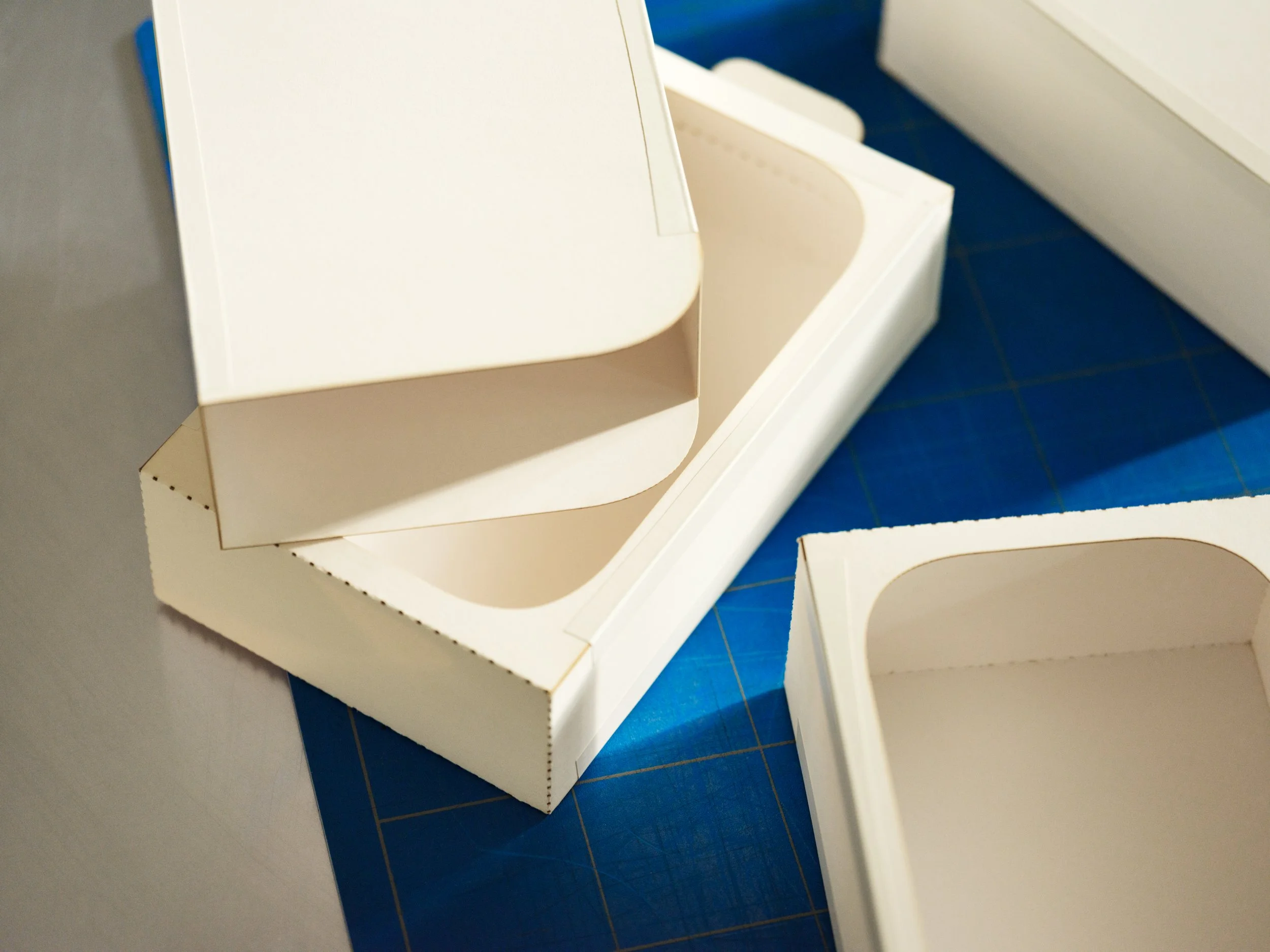Unassembled white cardboard or foam boxes on a blue cutting mat with grid lines.