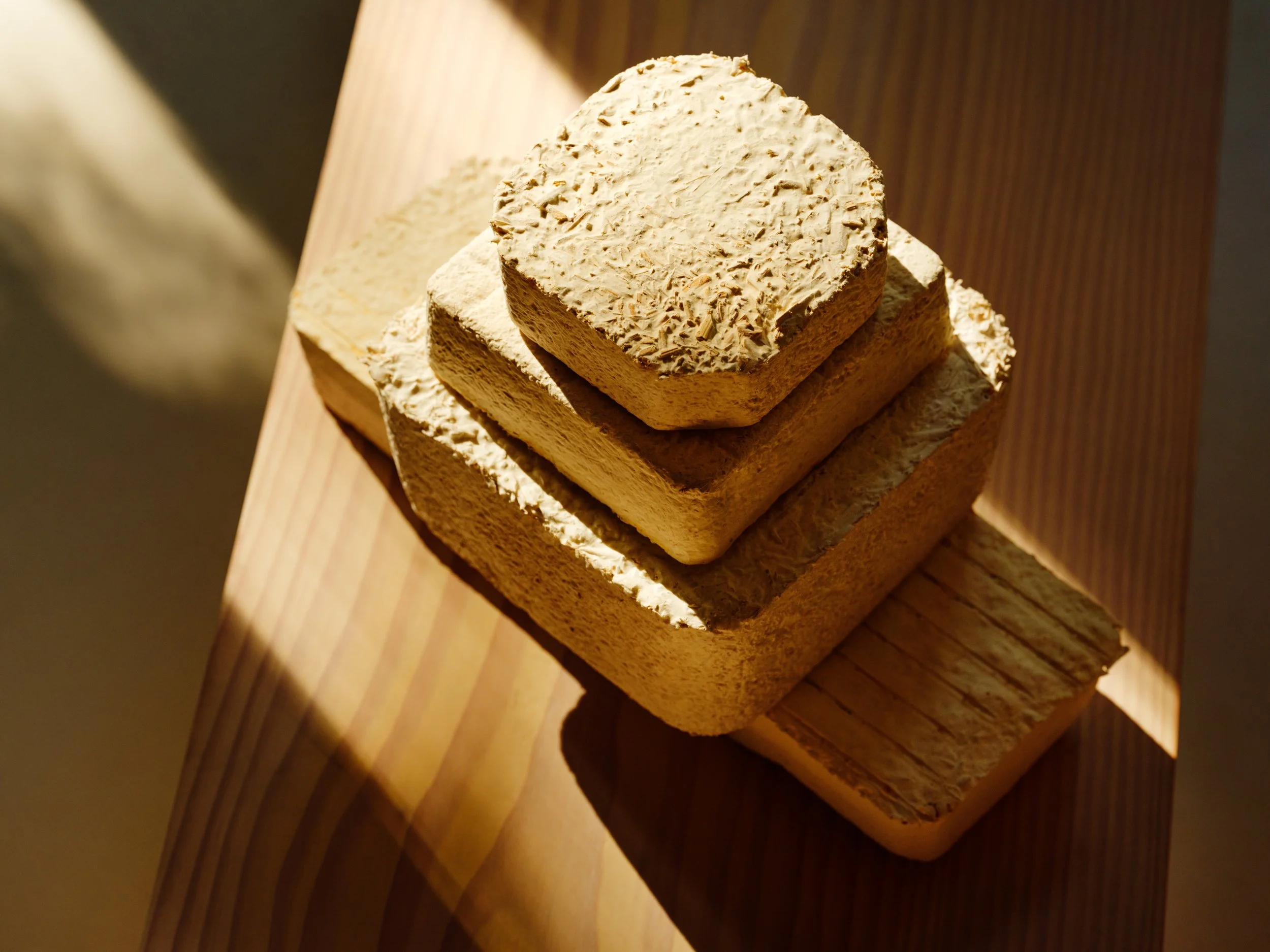Stack of four beige, square-shaped bricks with rough, textured surfaces placed on a wooden surface, illuminated by warm light.