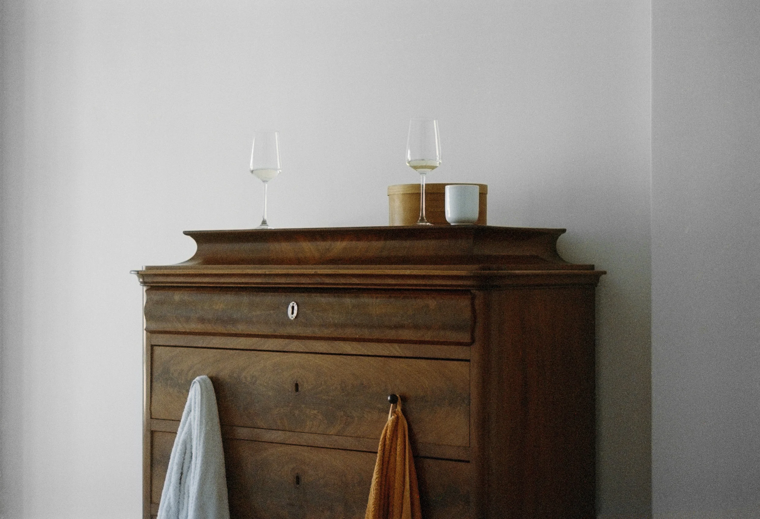 A wooden dresser with two glasses of white wine, a white mug, and a wooden box on top, against a plain light-colored wall.