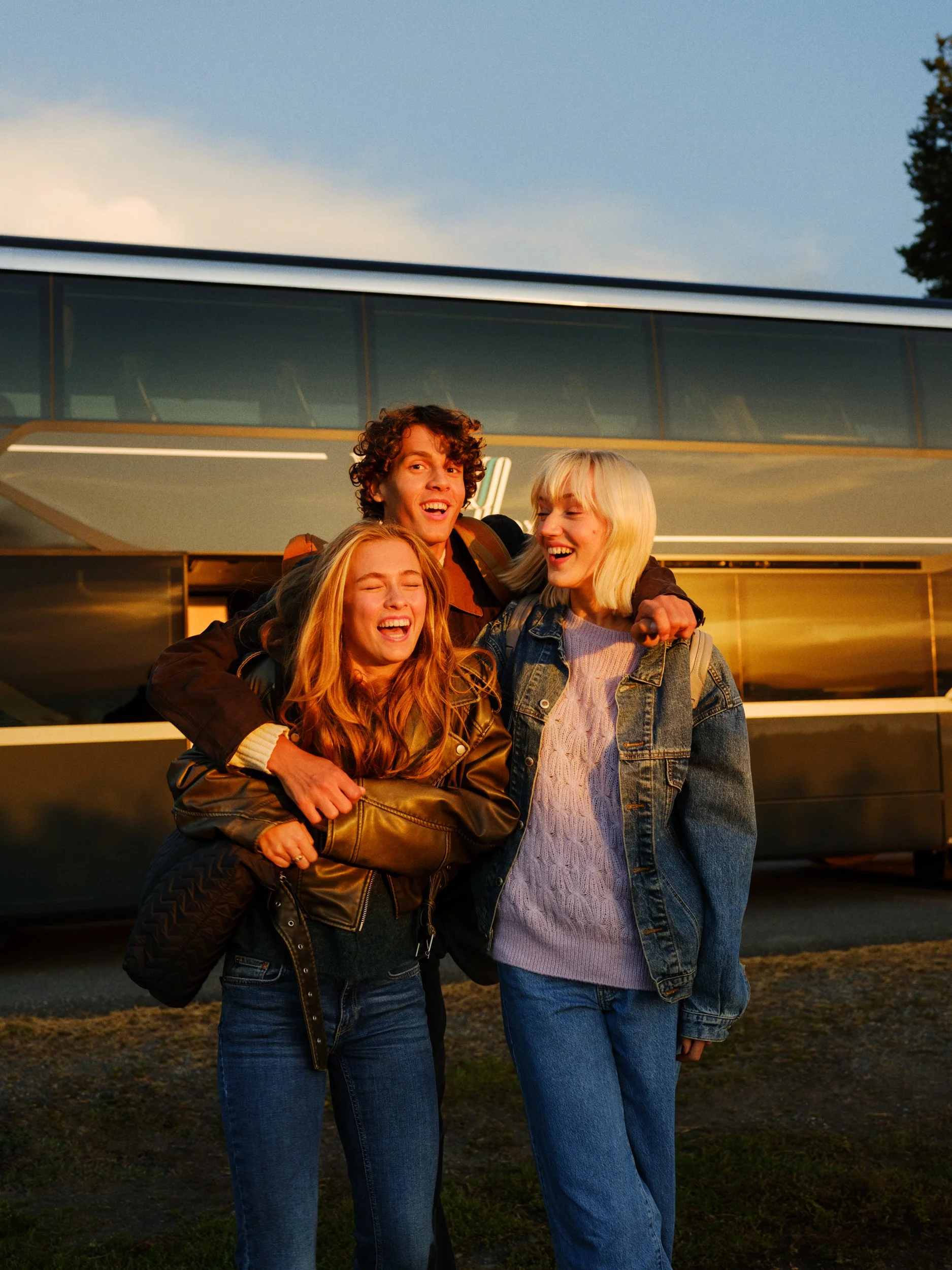 Three young friends, two women and one man, are smiling and hugging each other outside during sunset, with a modern building in the background.