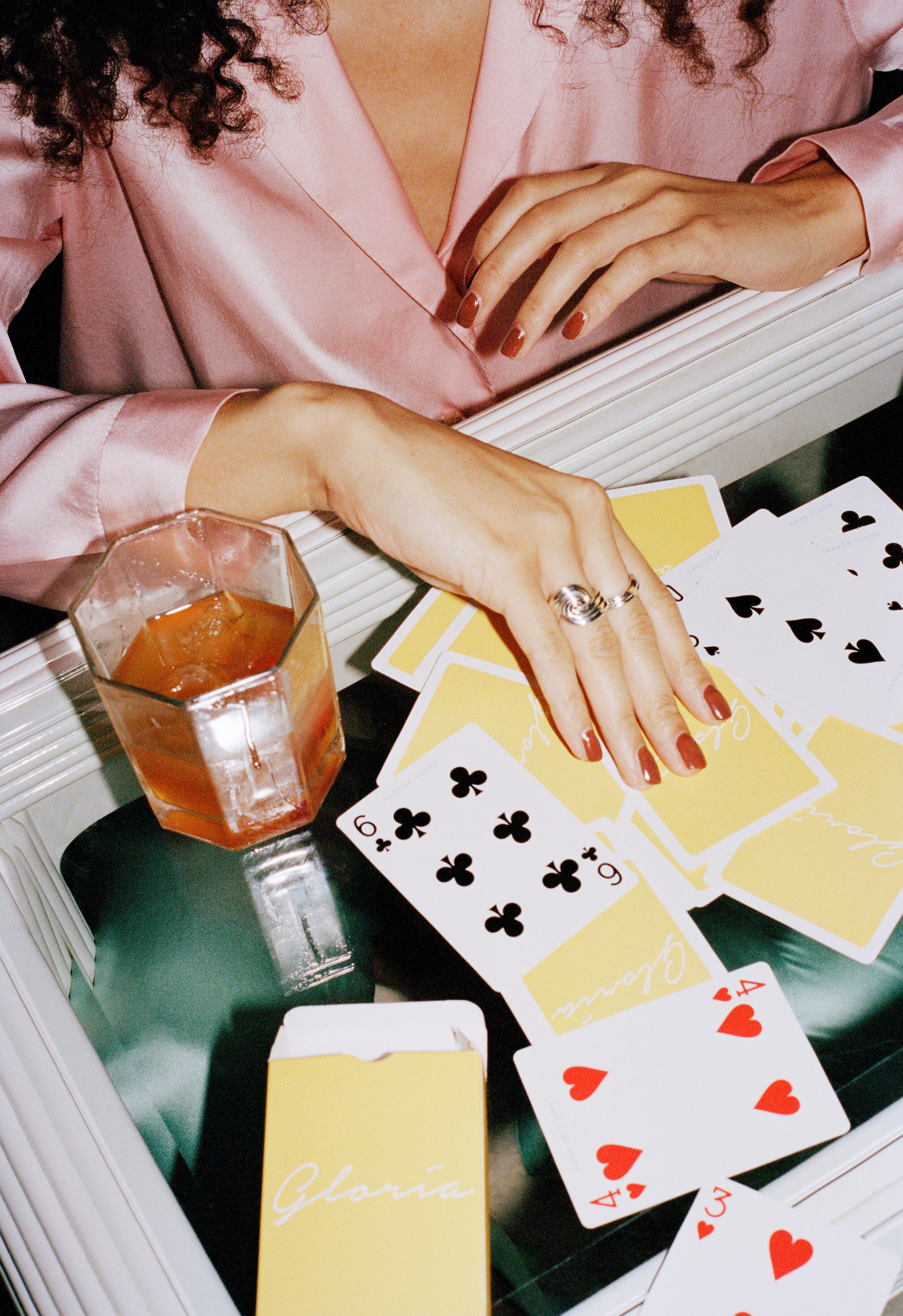 A woman wearing a pink satin blouse is playing cards on a black table. There is a glass of whiskey and ice in front of her, and her hands are placed on the cards, which include the six of clubs and the four of hearts. A yellow card box labeled 'Gloria' is also on the table.