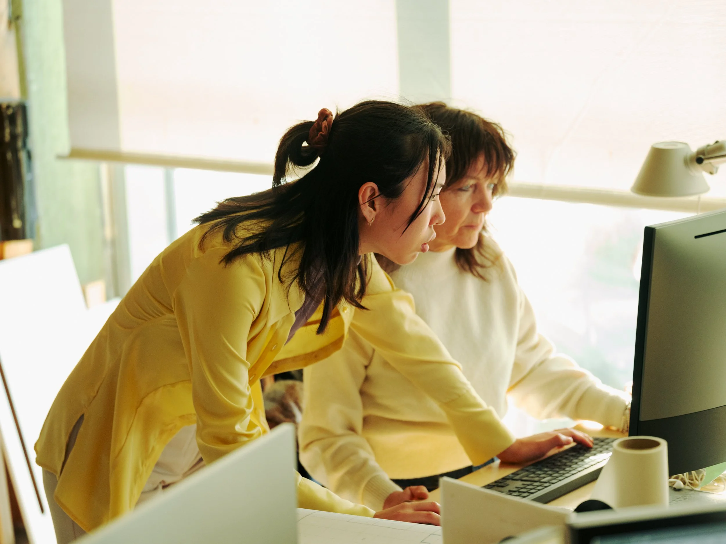Two women working together at a desk with a computer, in a bright office.