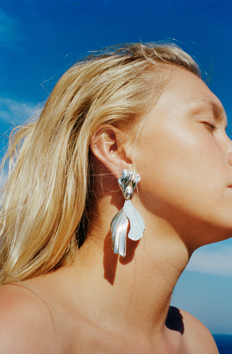 Close-up of a woman with blonde hair, wearing large silver earrings, with her eyes closed, against a clear blue sky.
