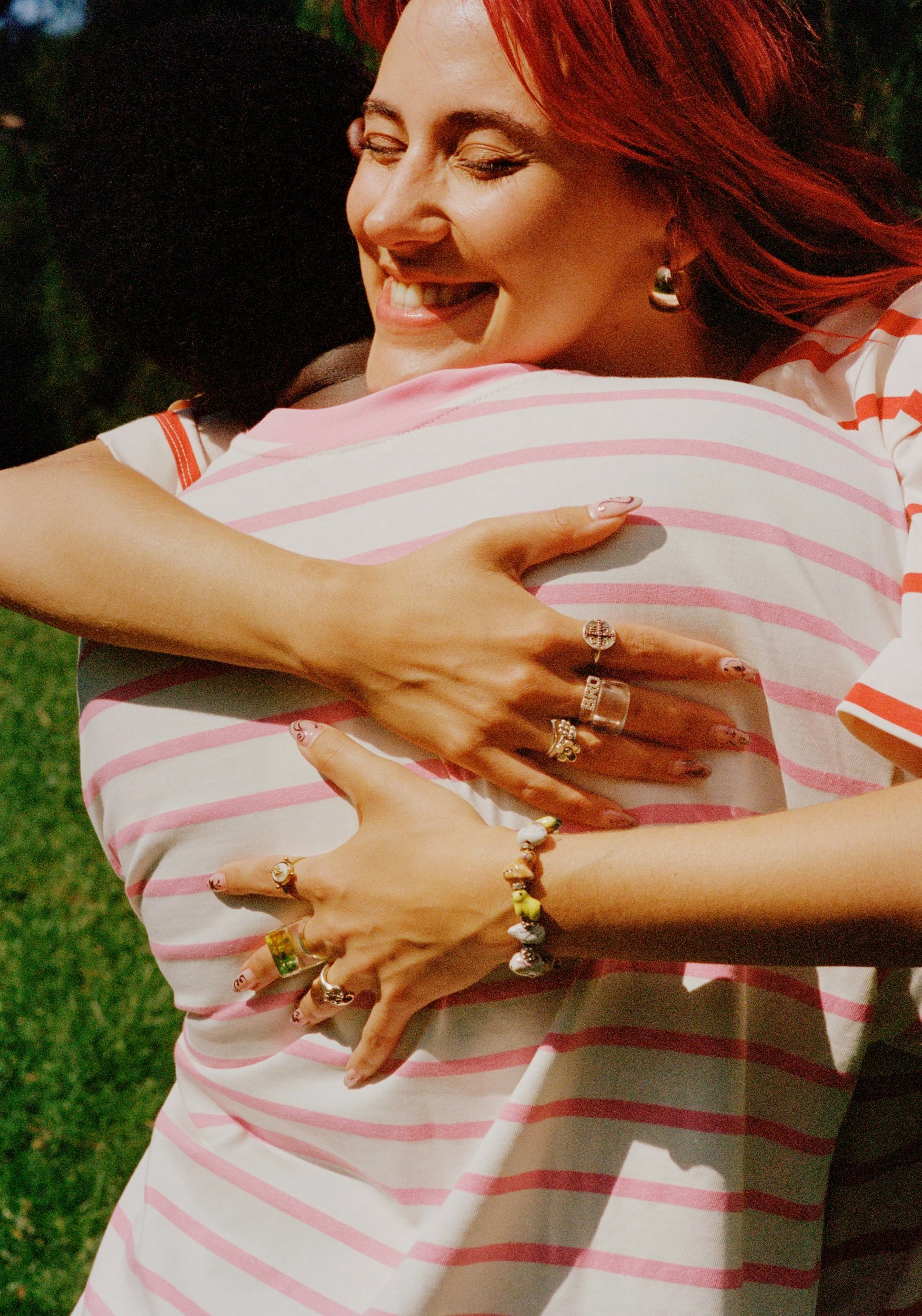 Two women hugging outdoors, one woman with red hair smiling joyfully, wearing jewelry and a striped shirt, the other woman with dark hair, wearing a similar striped shirt.