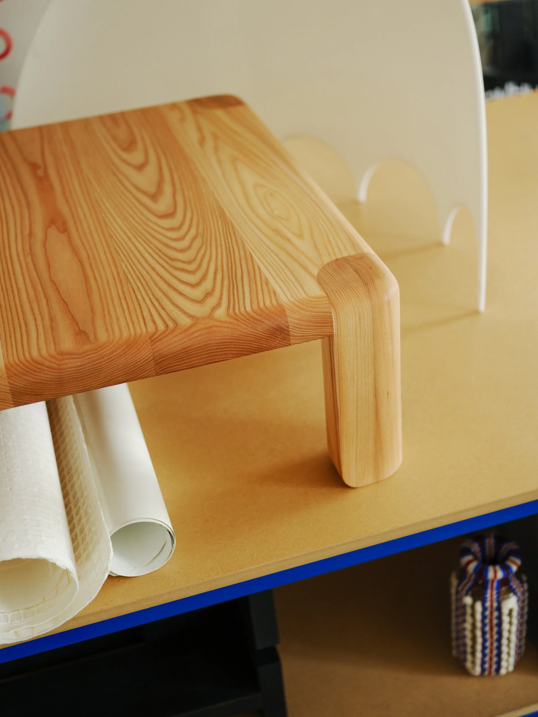 Close-up of a small wooden table with a rounded-edge top, placed on a yellow surface, with rolled-up paper and a small fabric bundle nearby.