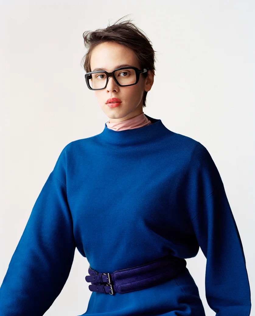A person with short, dark hair wearing large black glasses, a pink turtleneck under a blue oversized sweatshirt, and a purple belt.