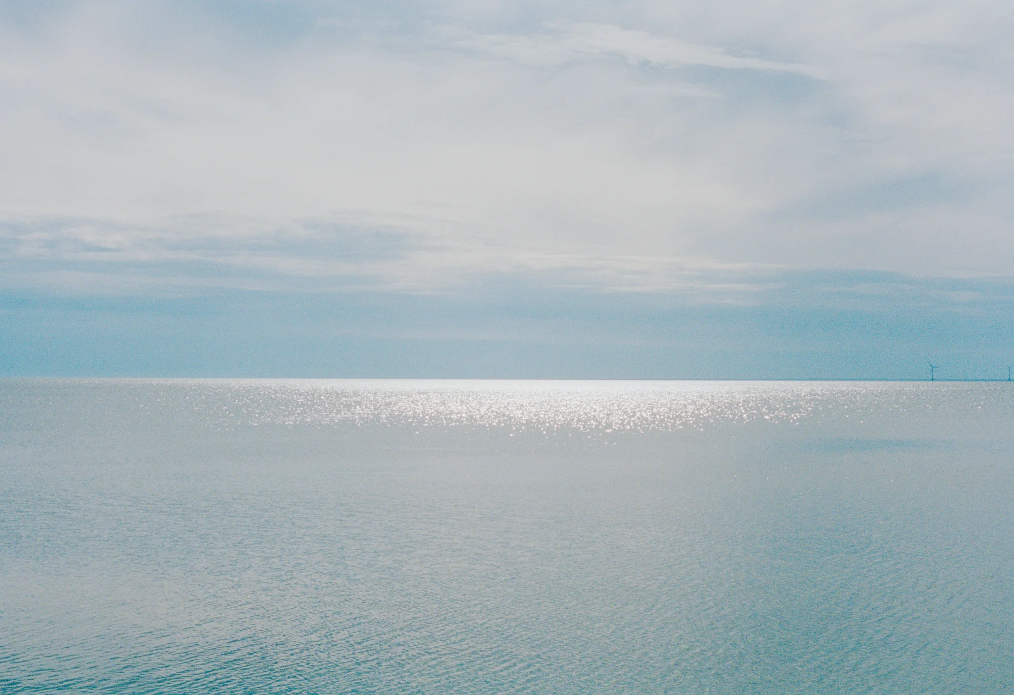 A calm ocean with sunlight reflecting on the surface and wind turbines on the horizon under a partly cloudy sky.