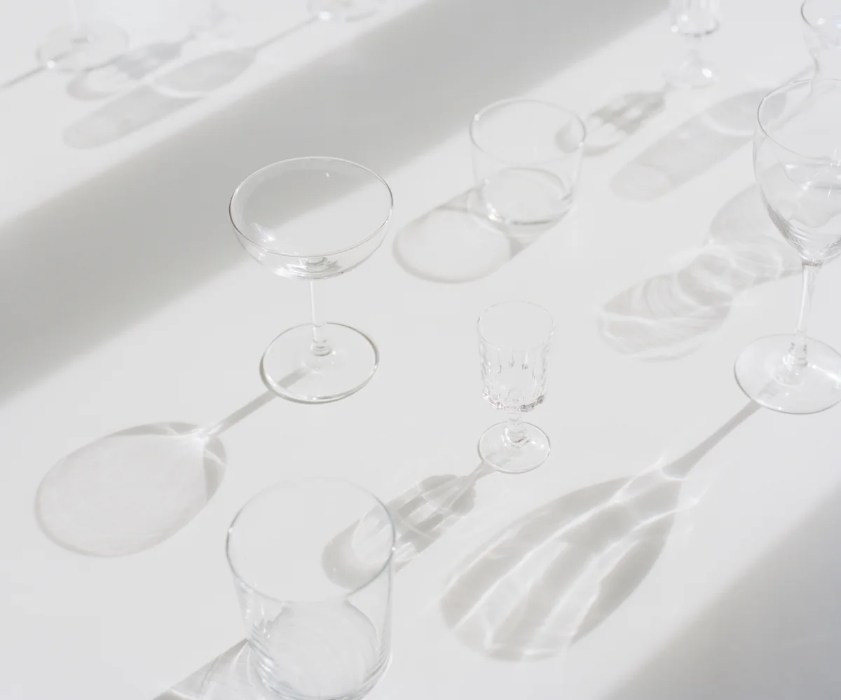 Several empty, clear glassware including wine glasses, a champagne flute, and a tumbler, casting shadows on a white surface.