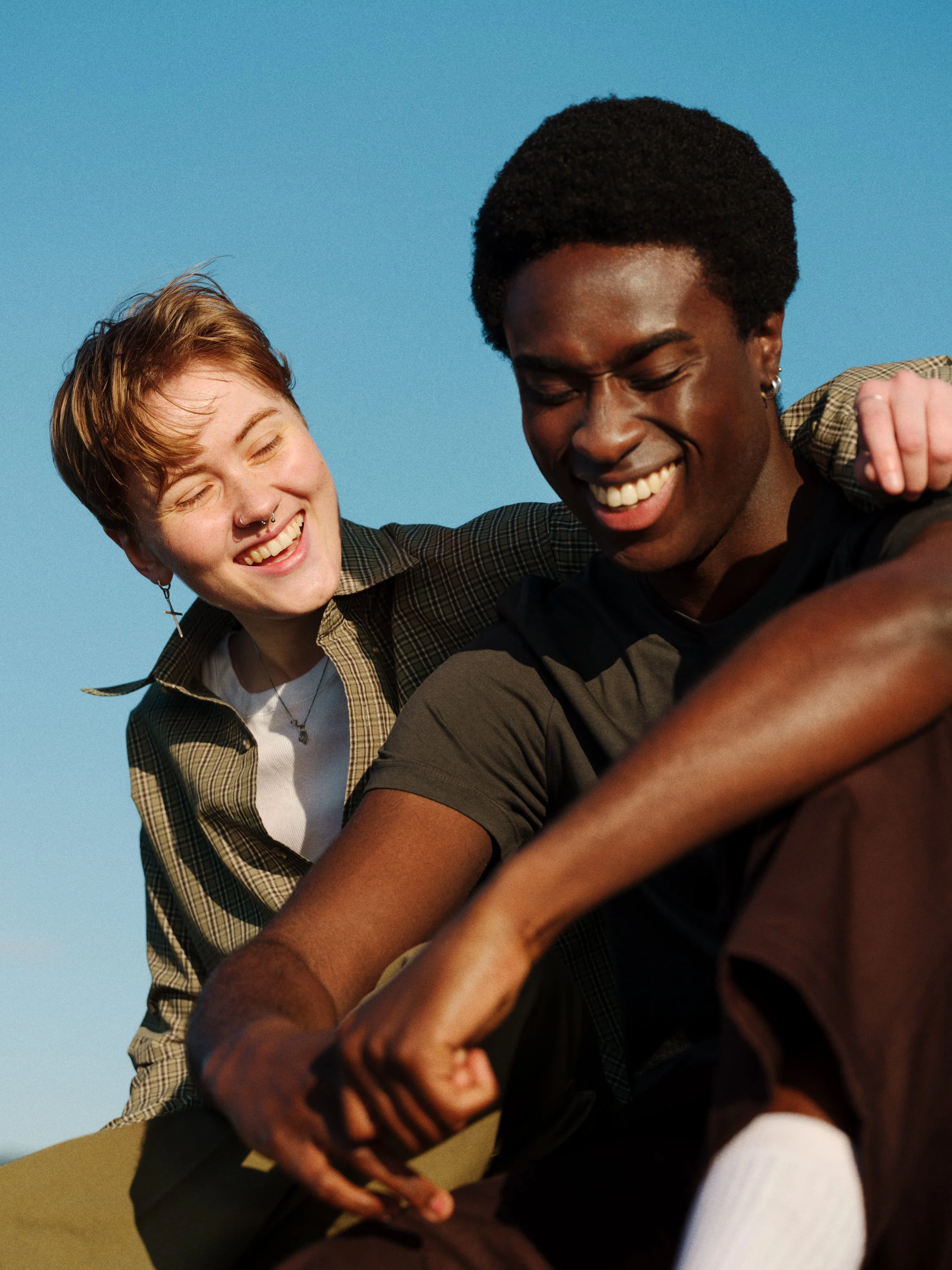 Two friends, a light-skinned woman with short, reddish hair and a dark-skinned man with an afro hairstyle, are smiling and playing together outdoors against a clear blue sky. They appear happy and are laughing.
