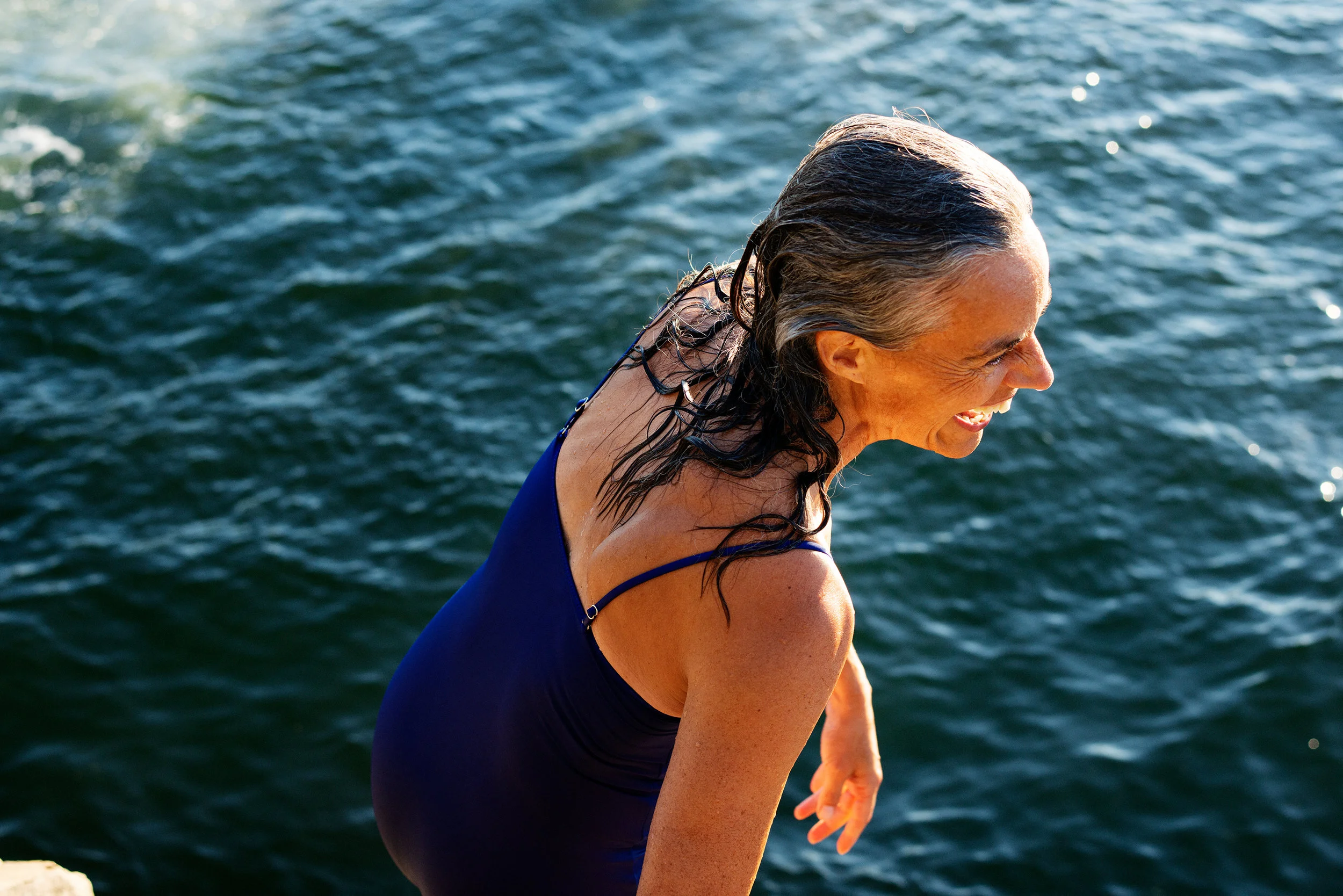 Older woman smiling and climbing out of a body of water, wearing a blue swimsuit with wet, dark hair.