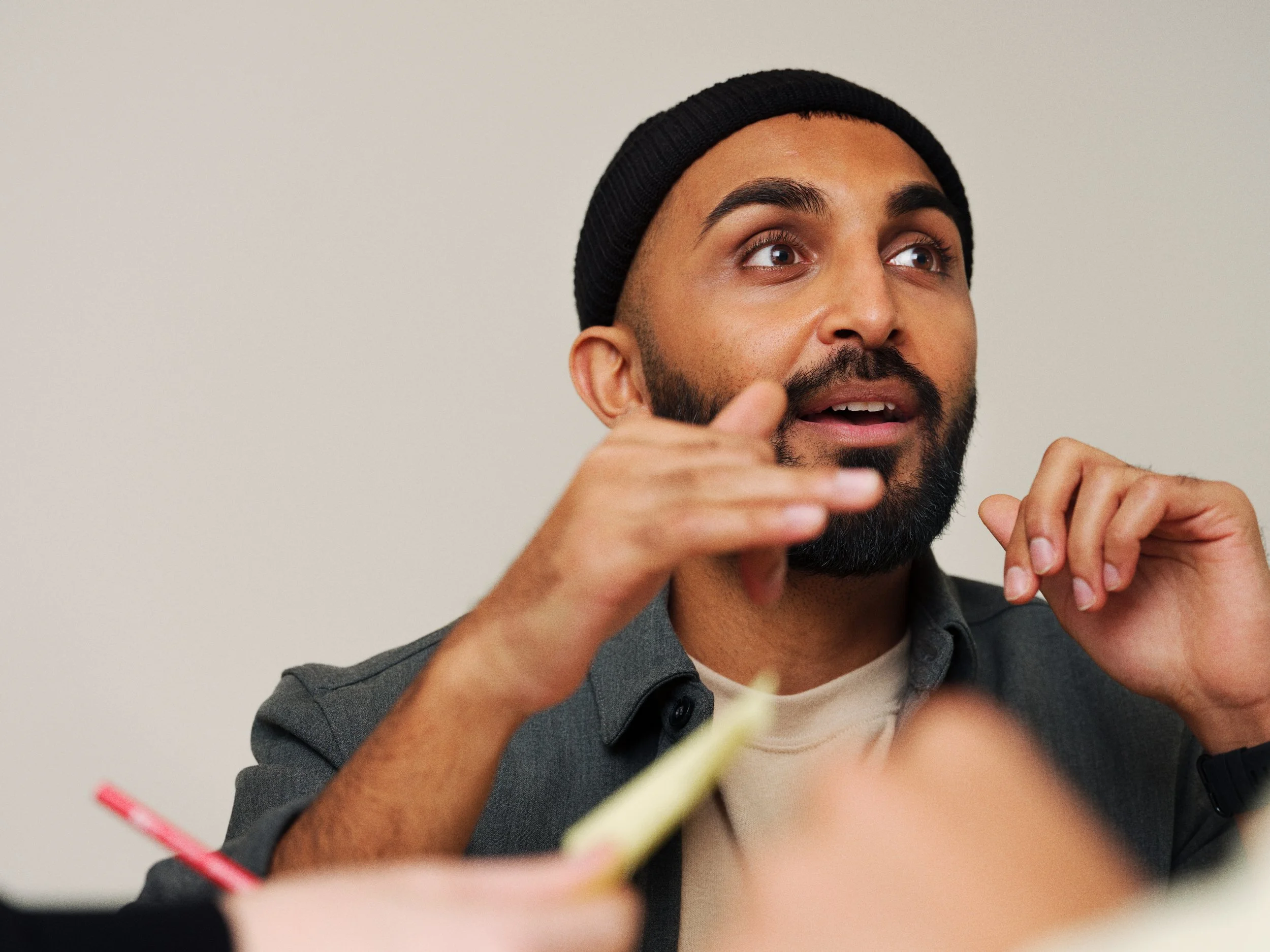 Man with a beard and black beanie talking and gesturing with his hands, sitting indoors.