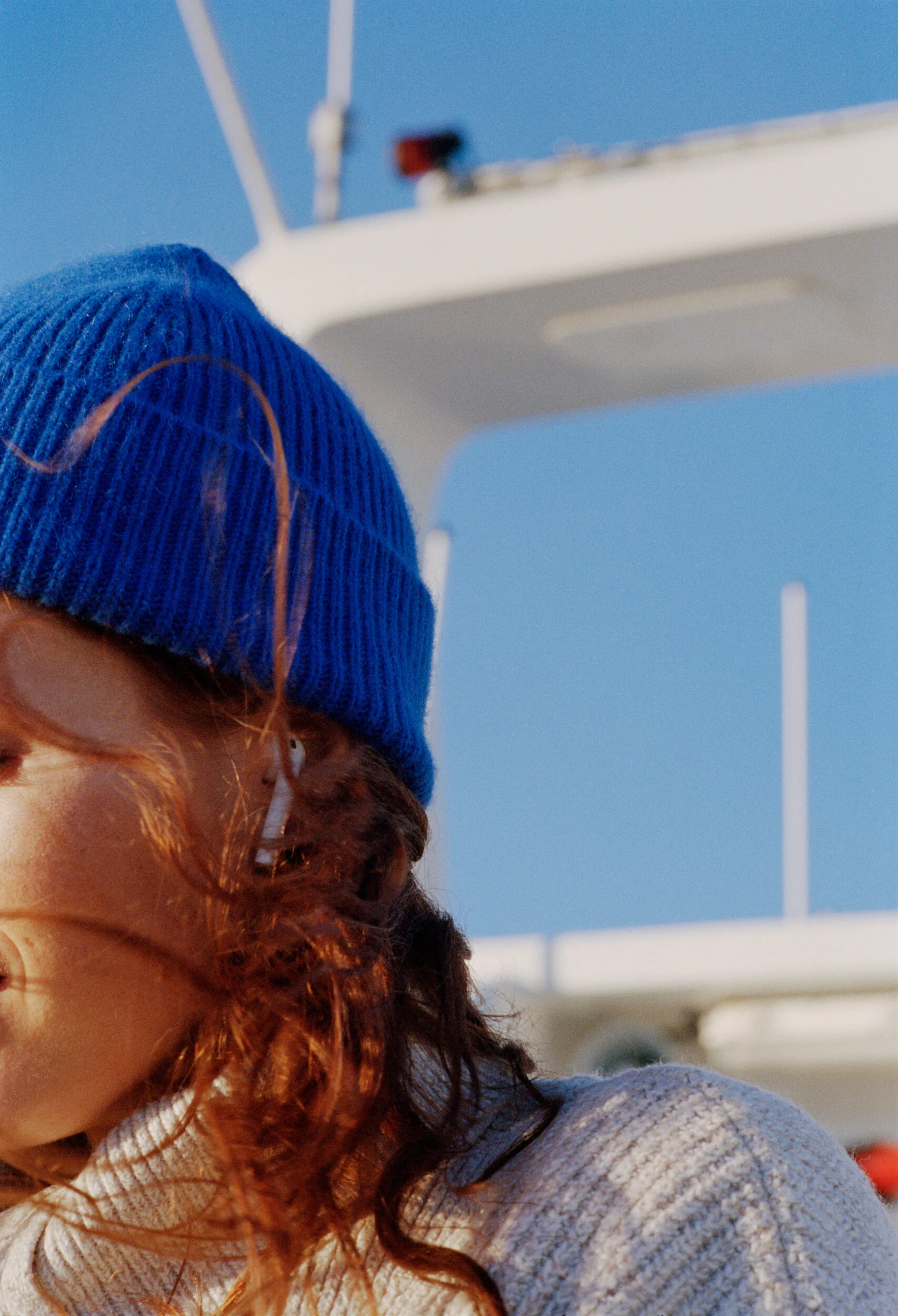 A person with curly red hair wearing a blue knit hat and white earbuds, standing in front of a large white boat or yacht against a clear blue sky.