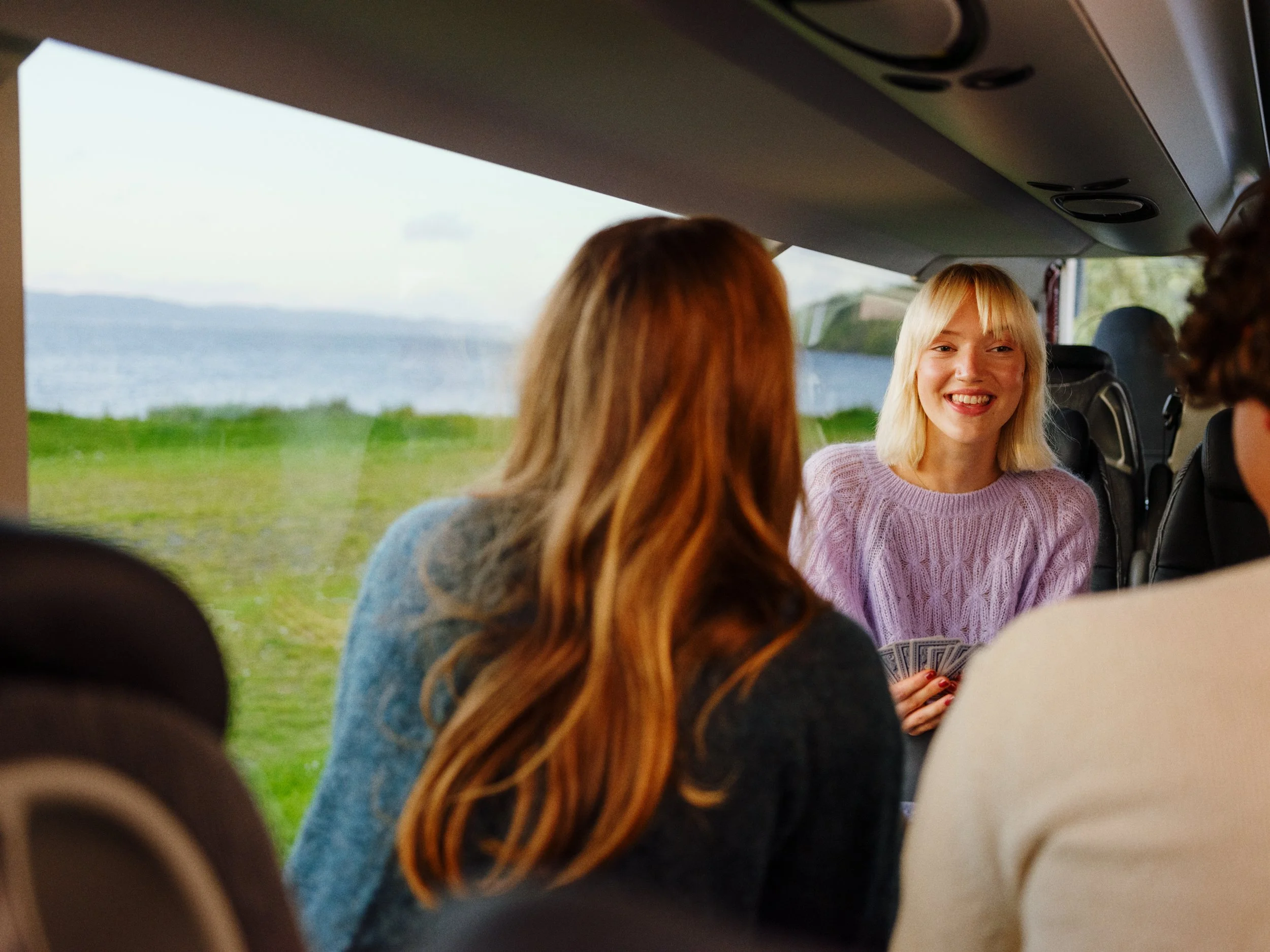 A group of friends sitting inside a bus or van, with a scenic view of water and green grass outside the window. One woman with blonde hair and a lavender sweater is smiling and holding playing cards, engaging with her friends.